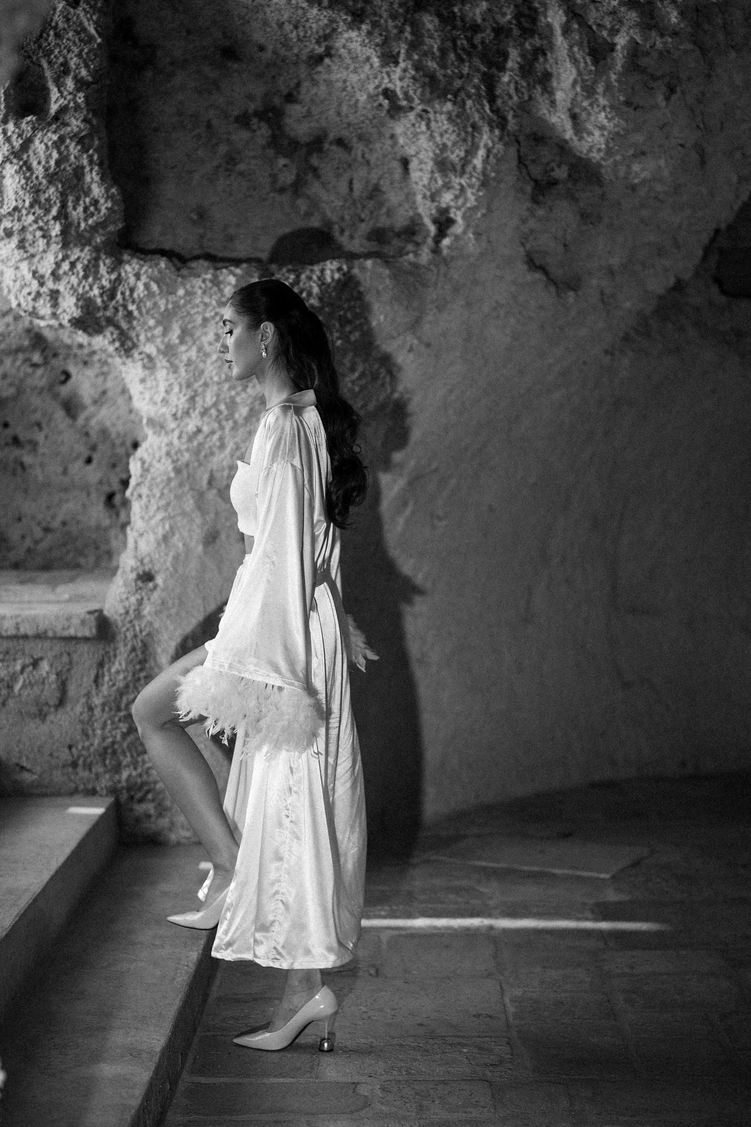 Black and white photo of a woman in a flowing white feathered robe and heels stepping up stone stairs in a rocky cave.