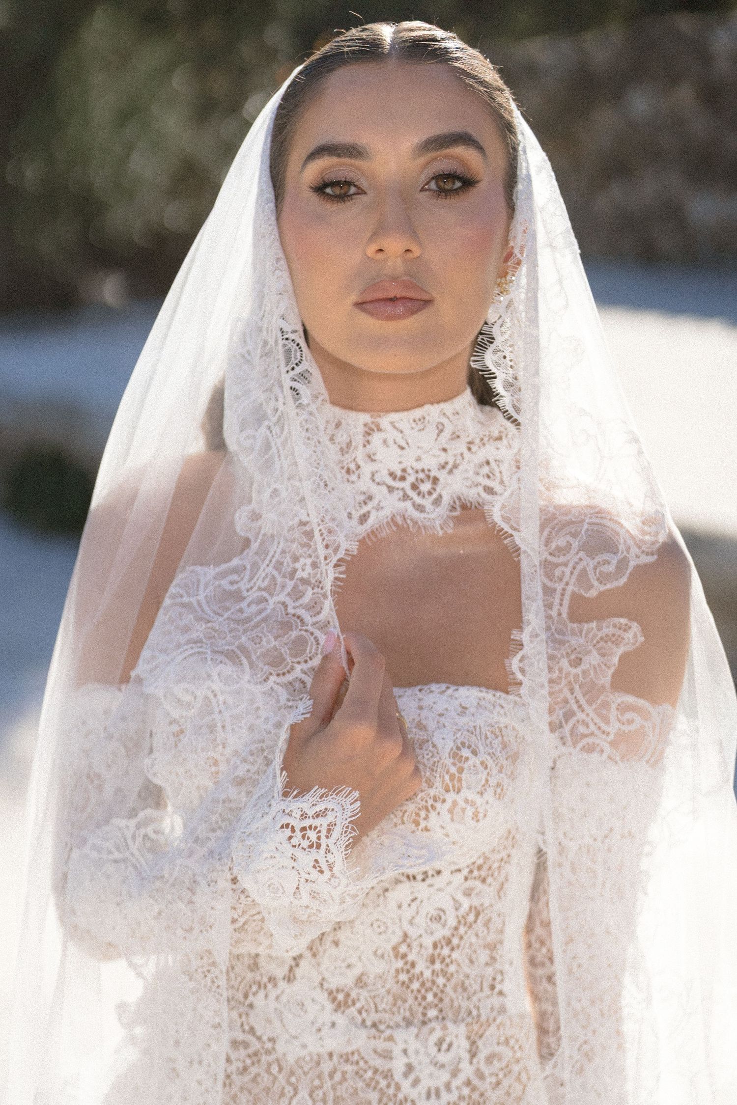 Close-up portrait of a bride in a lace wedding gown with a long lace-trimmed veil, hand resting near her collarbone, outdoors.