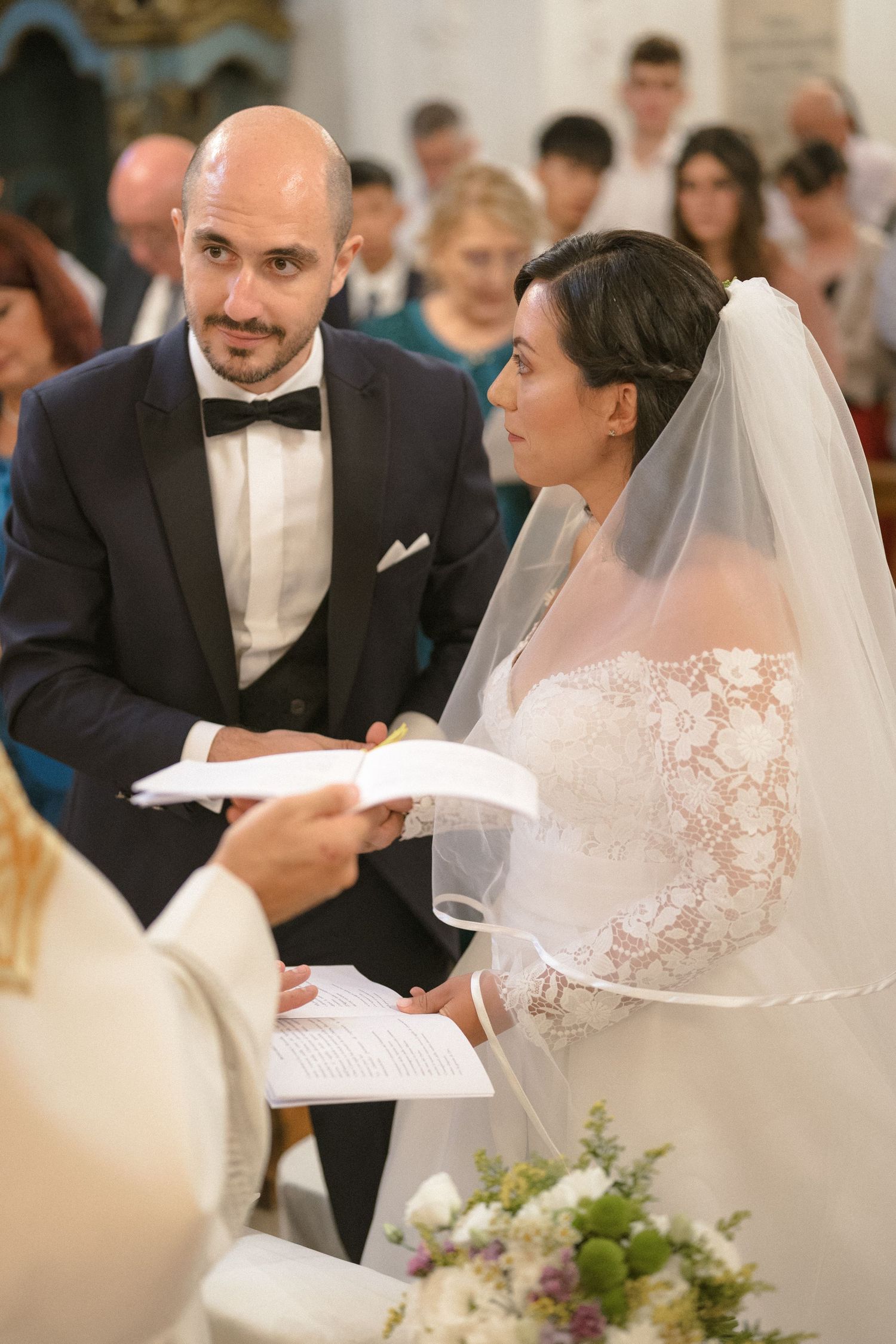 A bride and groom exchange vows during a traditional church wedding ceremony with guests in the background.