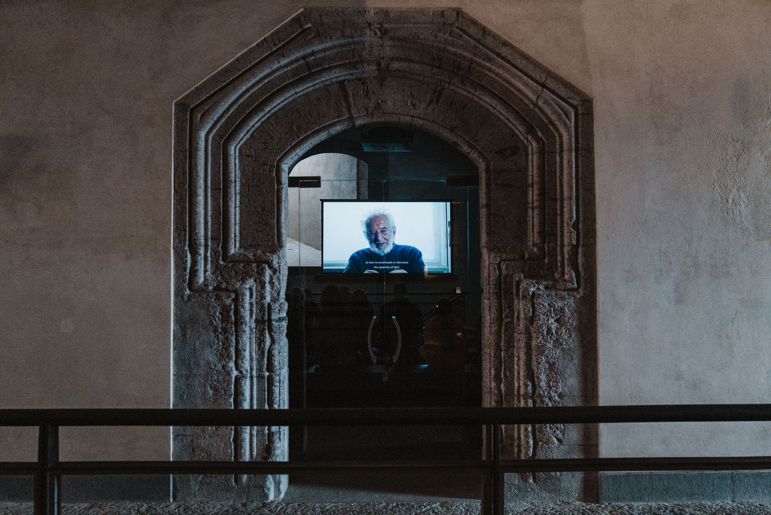 A television screen glows through an arched stone doorway in a dark corridor.