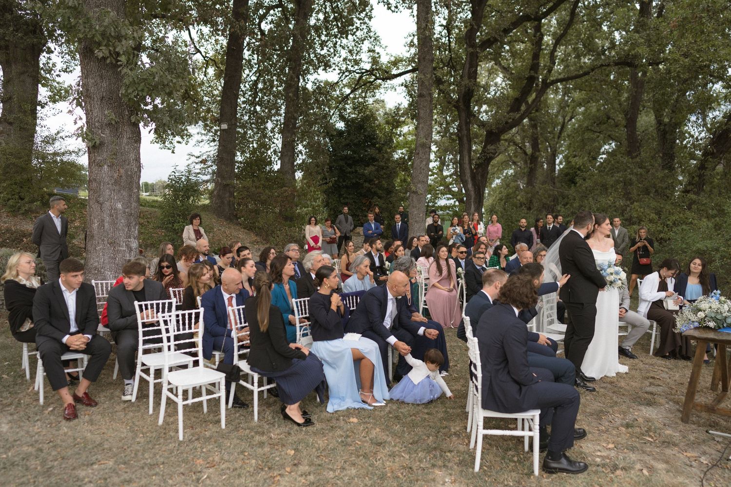 Wide view of wedding guests seated at an outdoor ceremony among trees.