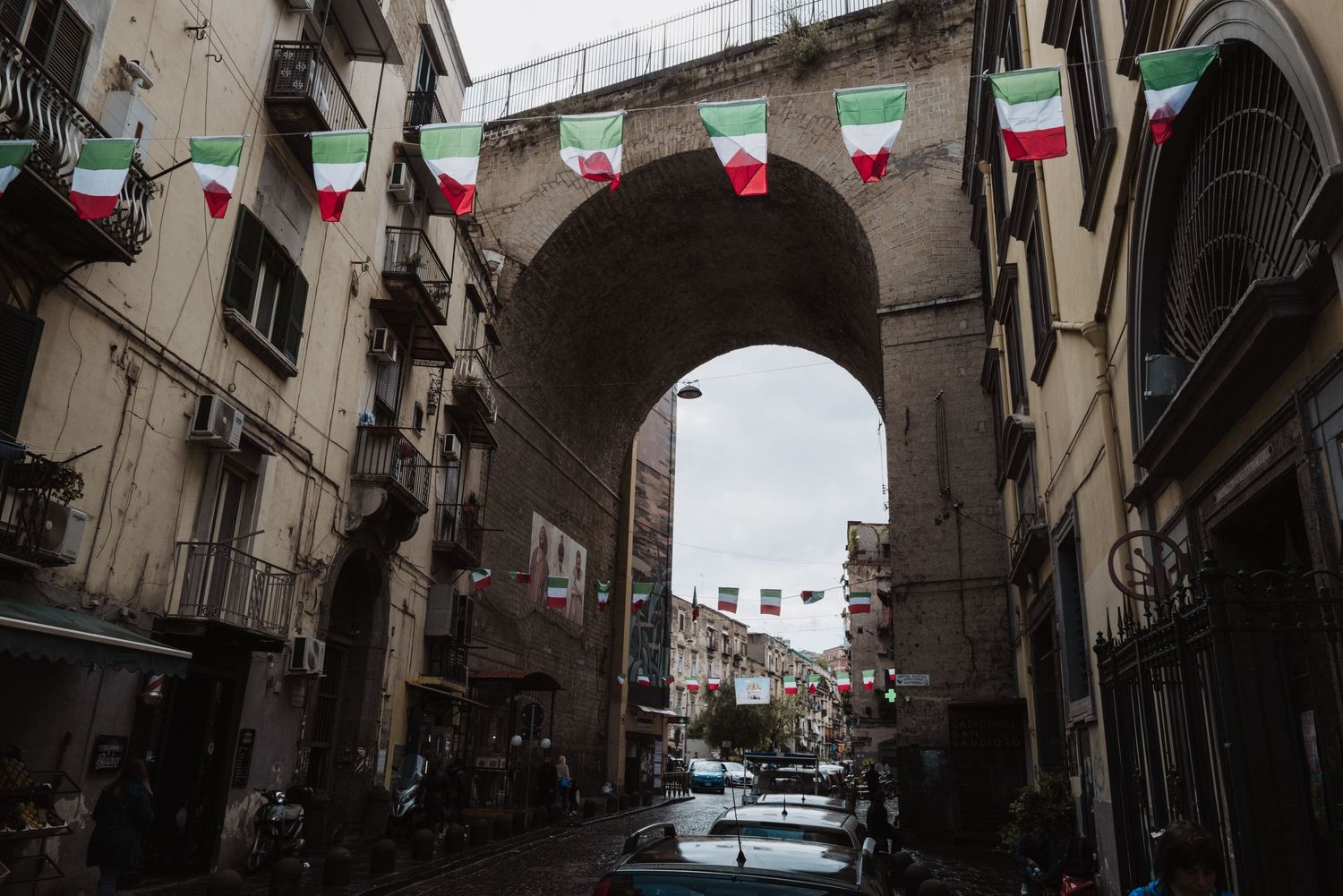 Italian flags hang over a narrow street viewed through an arched passageway in an urban setting.