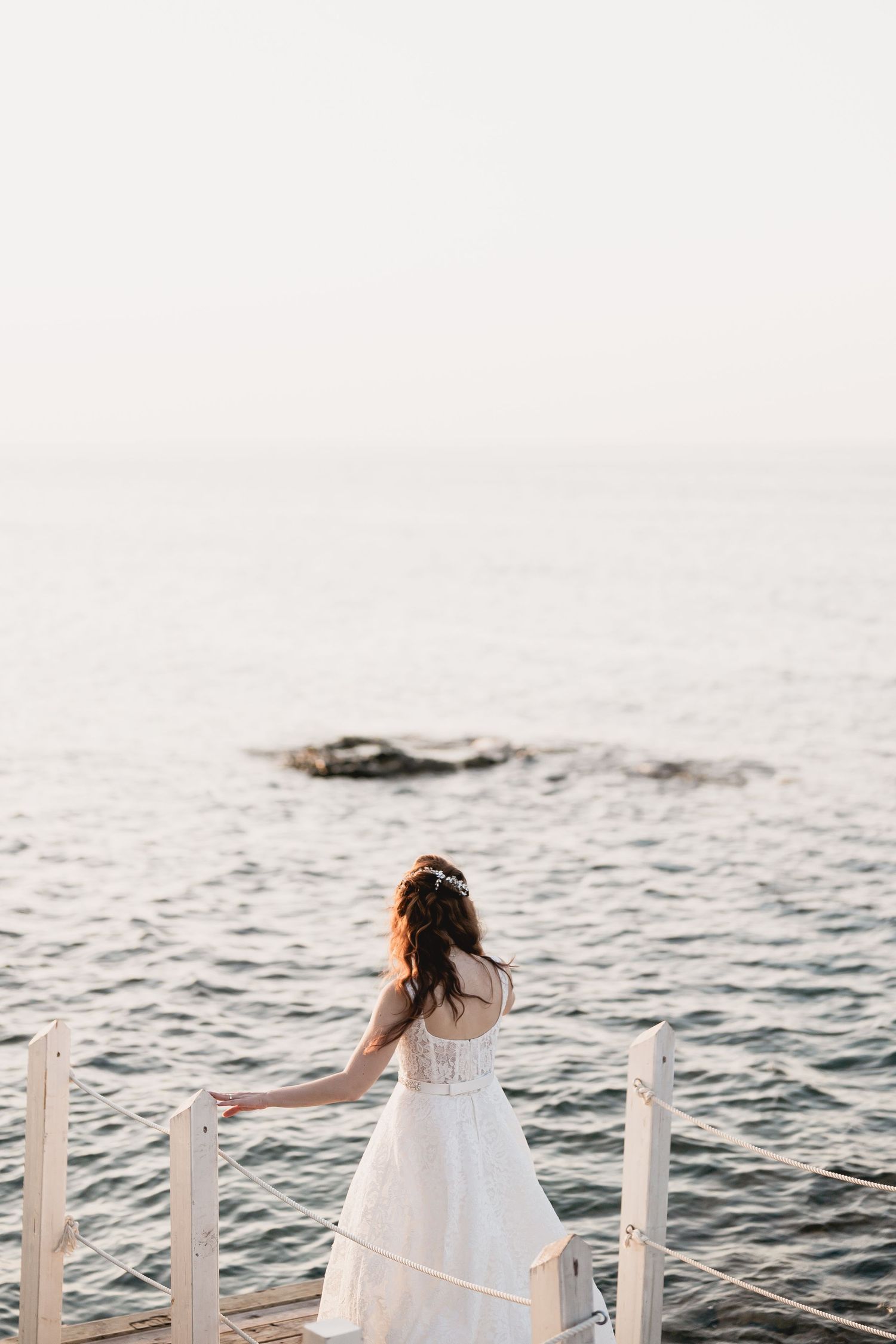 Bride stands on seaside deck overlooking ocean waves in dreamy wedding portrait.