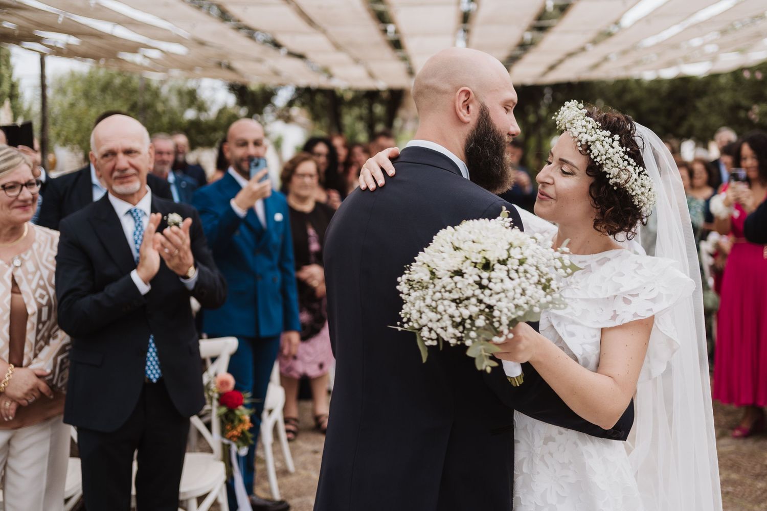Garden wedding ceremony with guests celebrating beneath a white canopy as a couple embraces with baby's breath bouquet.