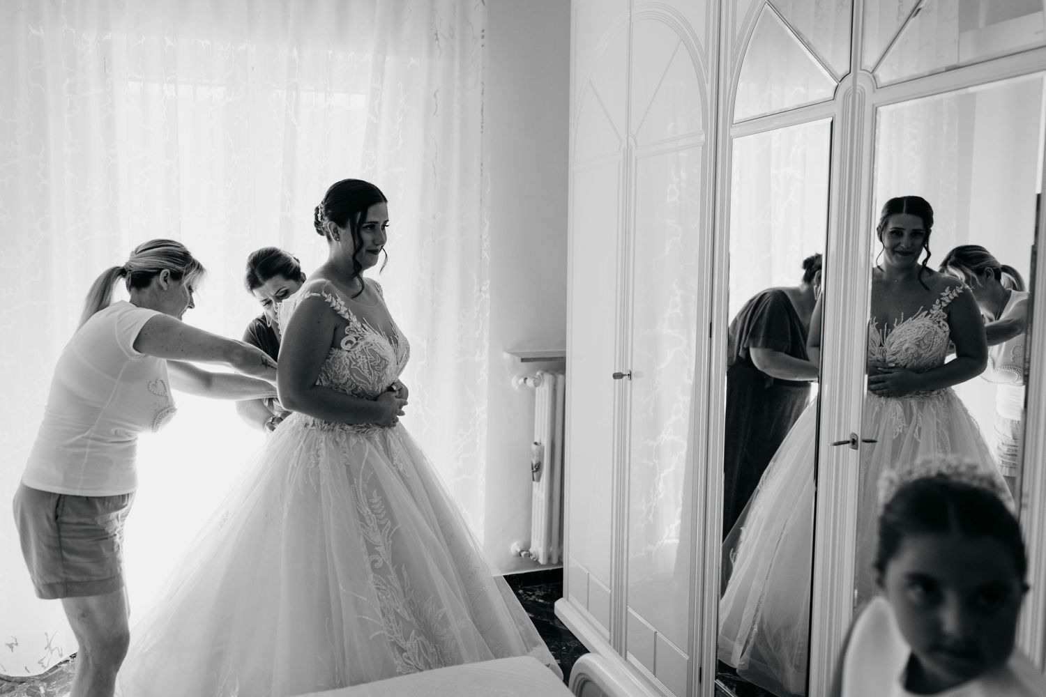 Black and white photo capturing wedding preparations as a bride gets ready in a bright room with a mirror reflection.
