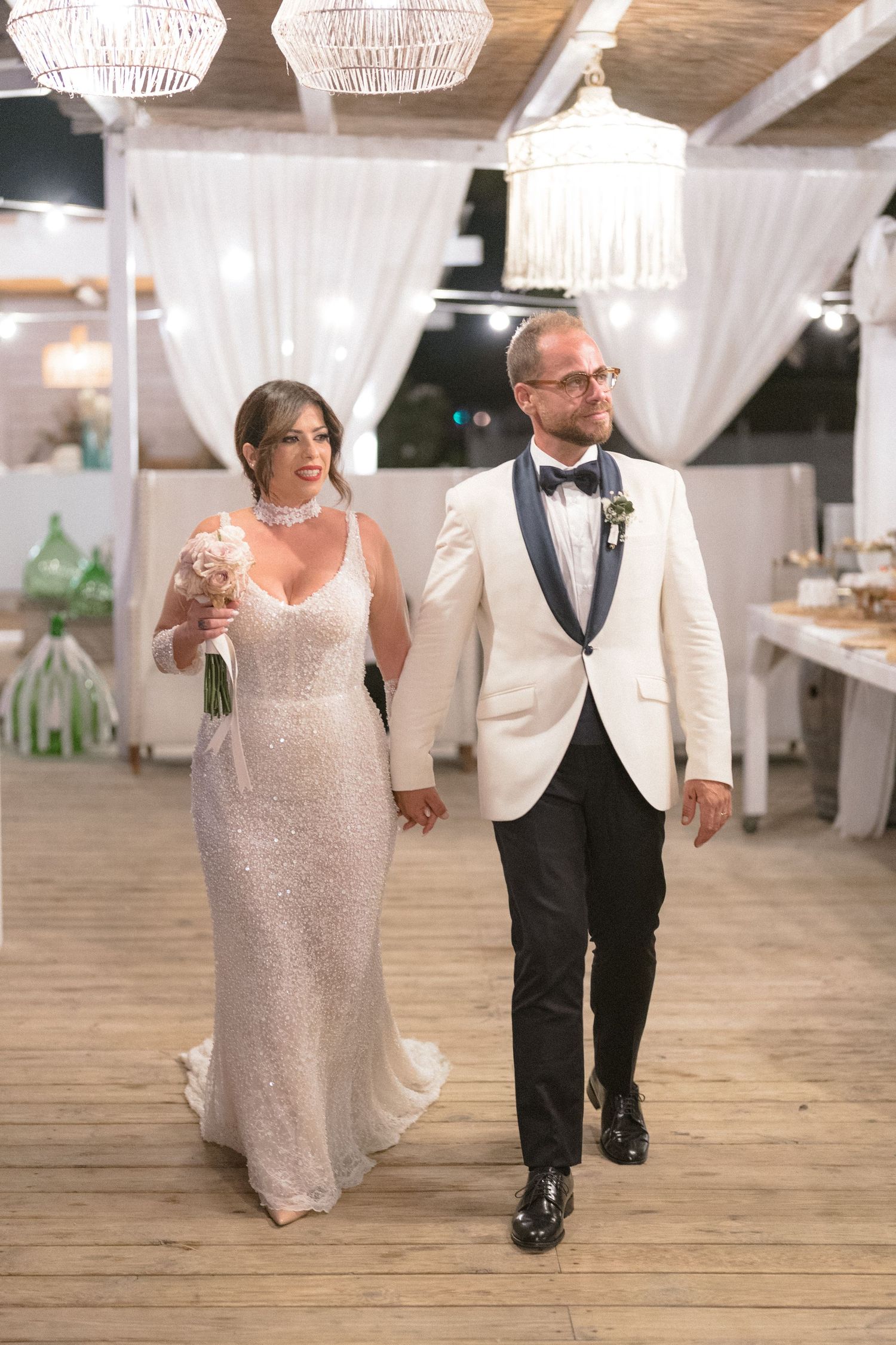 A wedding couple walks hand in hand through an elegantly decorated reception venue with crystal chandeliers.