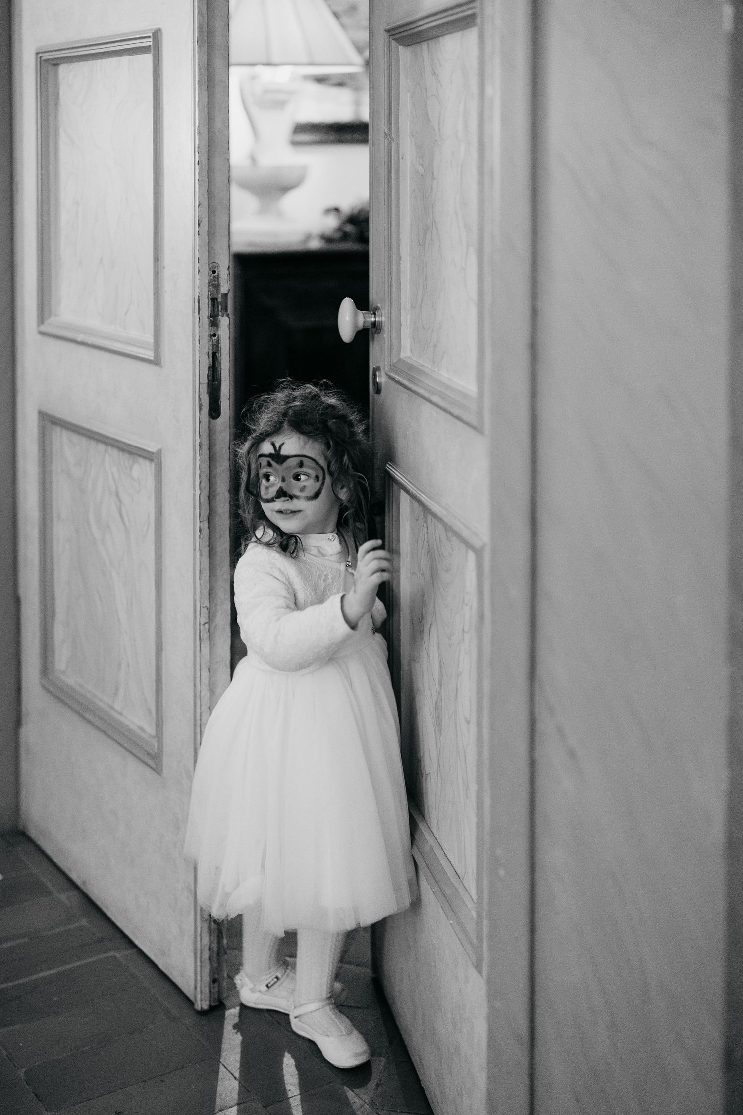 Adorable flower girl in white dress holding a masquerade mask while peeking through wooden doors.