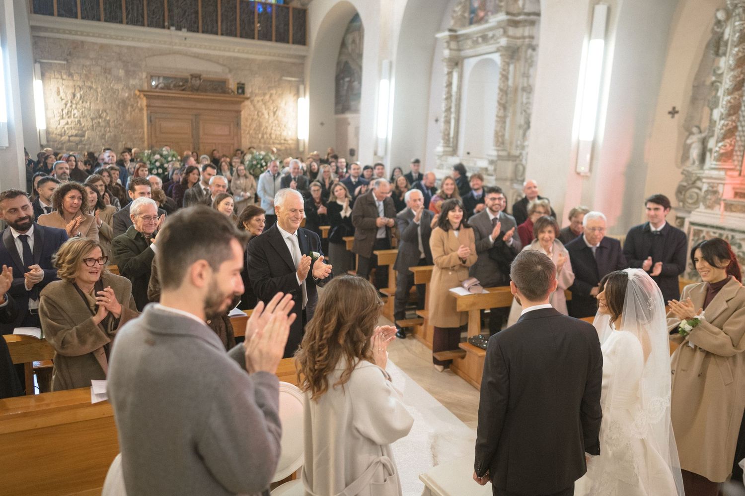 Wedding guests applaud as bride and groom stand at altar inside ornate church ceremony.