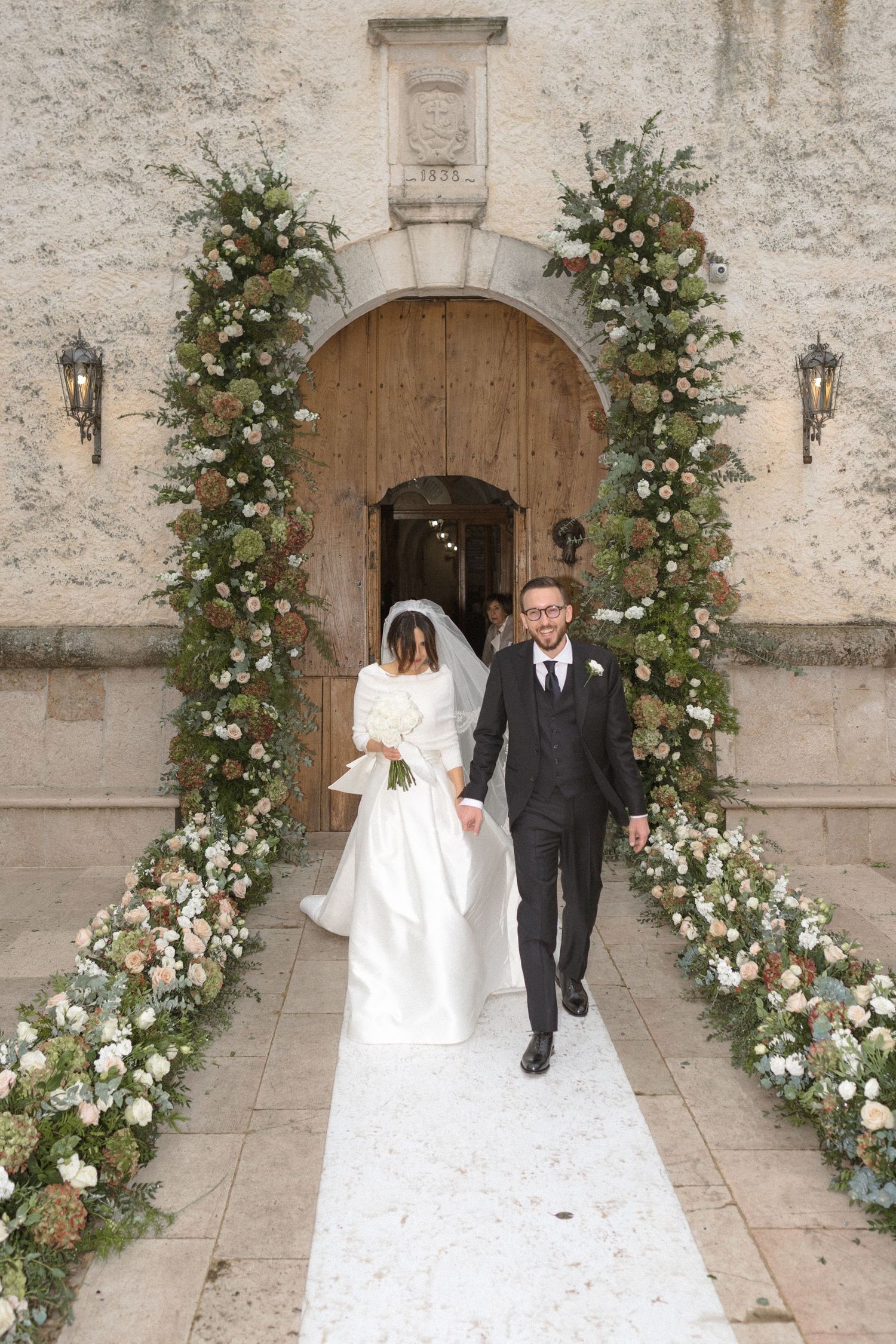 Bride and groom walk down floral-lined aisle outside stone church entrance decorated with lush greenery and white flowers.