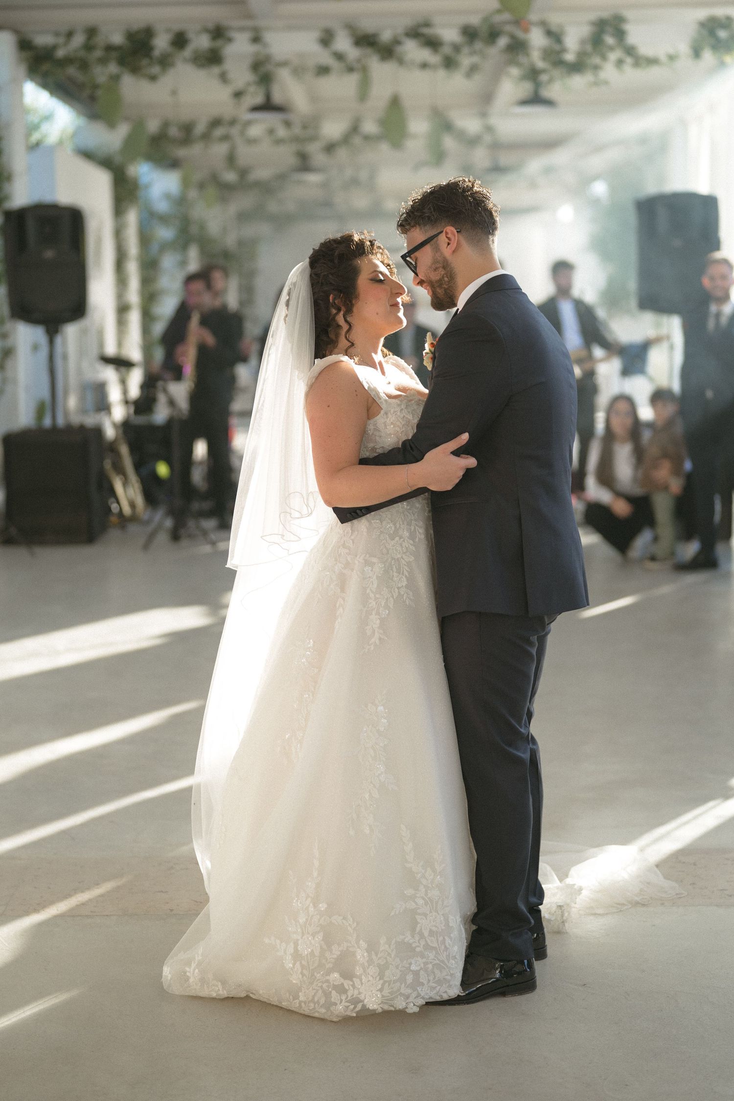 Bride and groom share first dance in sunlit reception venue with live band performing behind them.