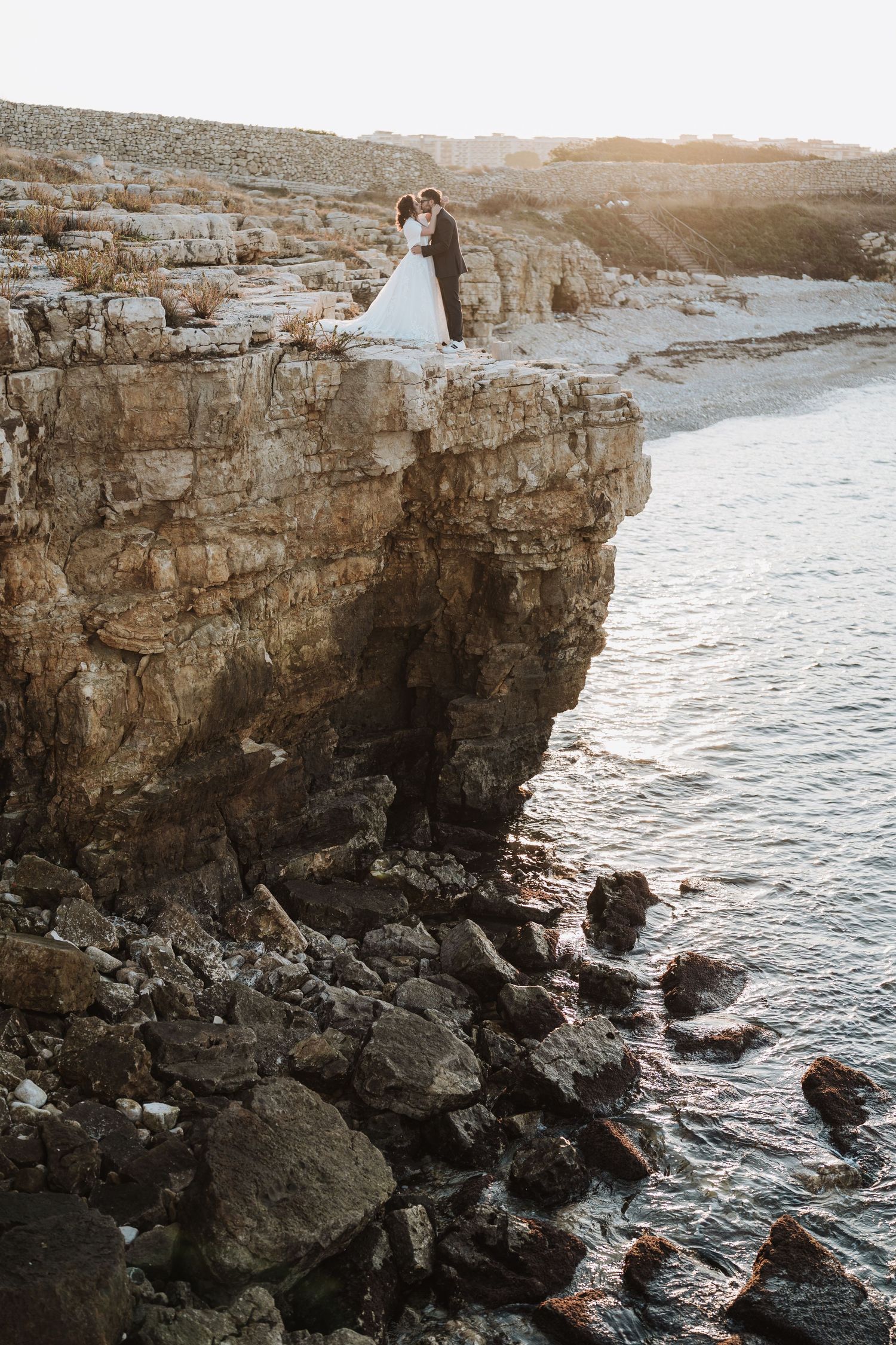 Bride and groom standing on rocky coastal cliffs at sunset, with ocean waves below in a romantic wedding photo.