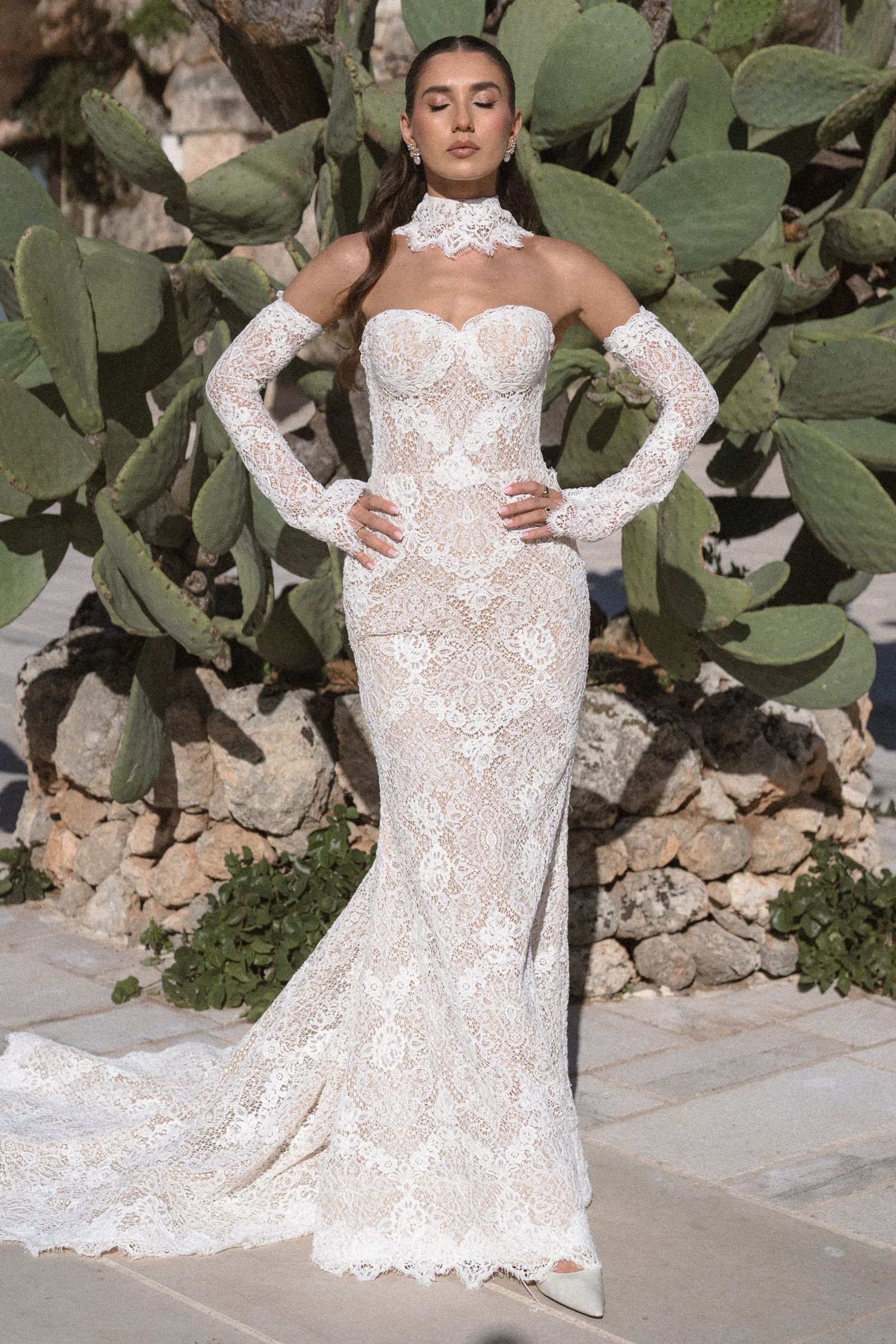 Bride in a fitted white lace mermaid wedding gown with a high collar and sheer sleeves stands before green prickly pear cacti.