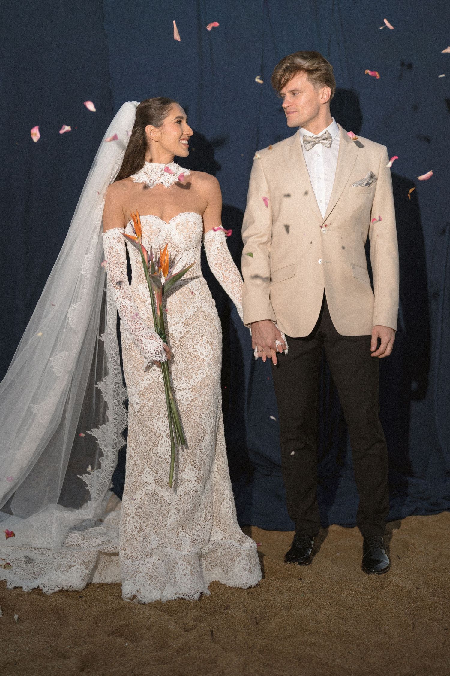 Smiling bride in a lace gown and veil holds a tropical bouquet and hands with her groom in a beige blazer as petals fall.