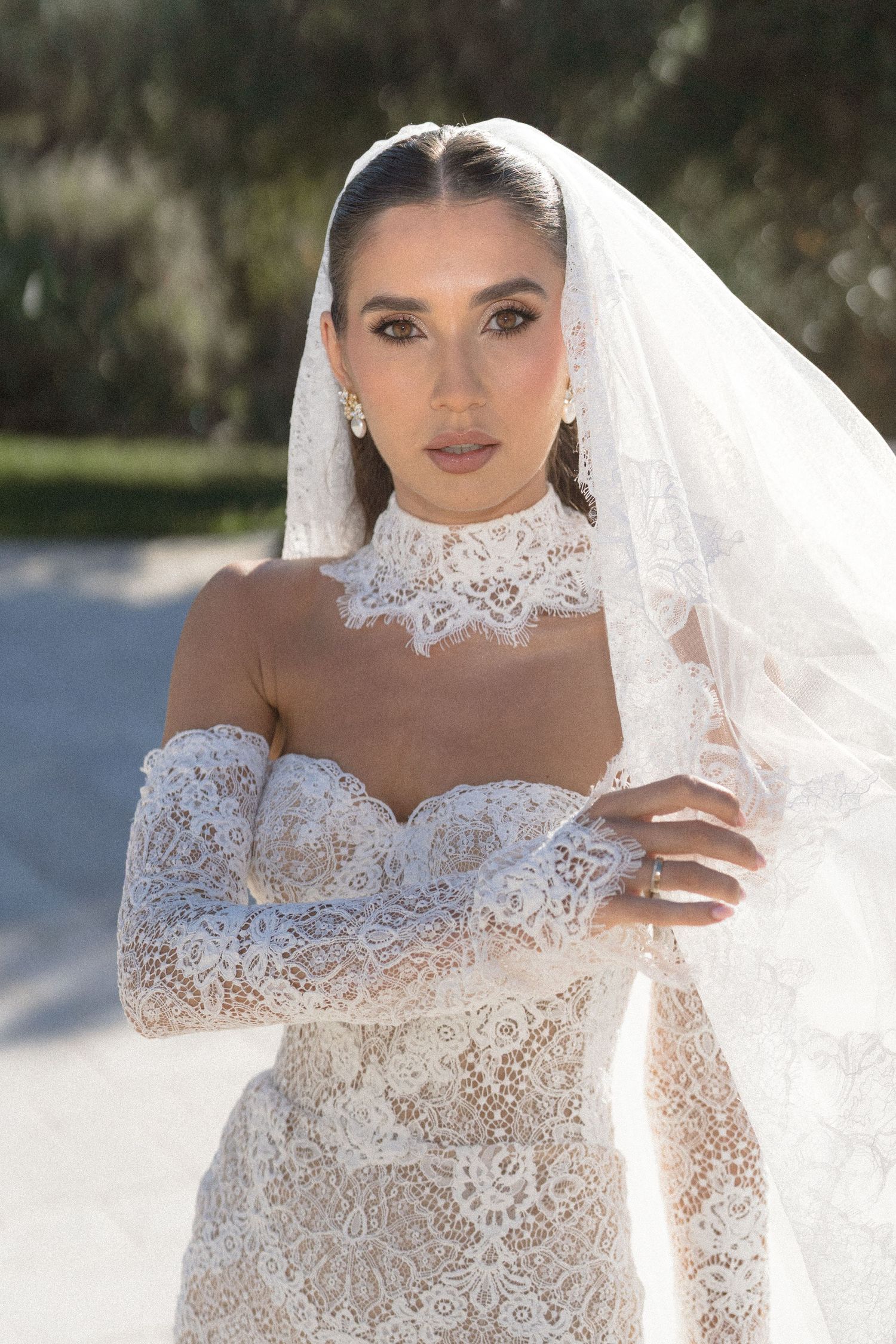 Bride facing the camera in a strapless lace wedding dress with off-shoulder lace sleeves, a high collar, and a long veil.