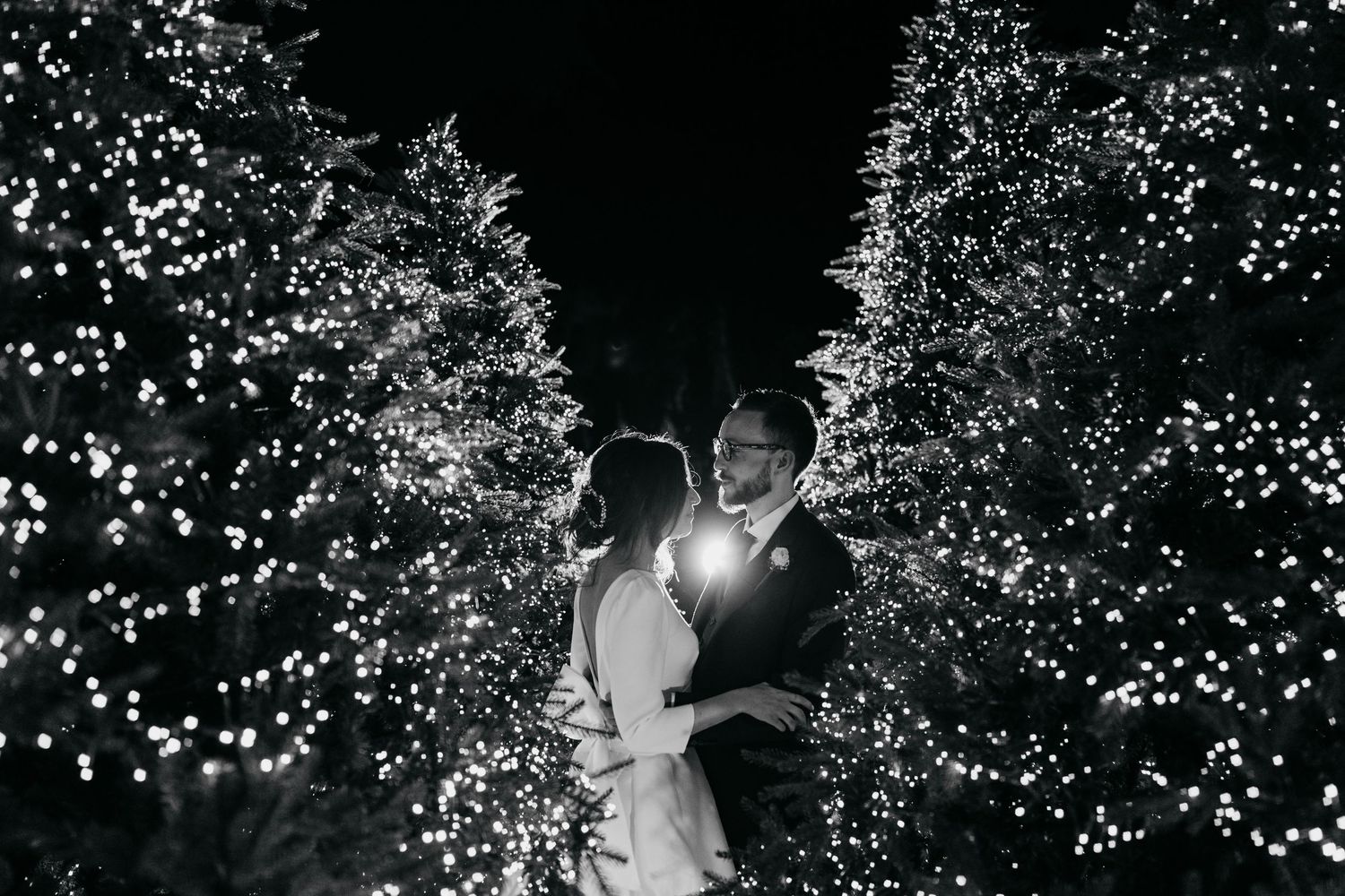 Black and white romantic wedding portrait of couple kissing among glowing light-wrapped trees at night.