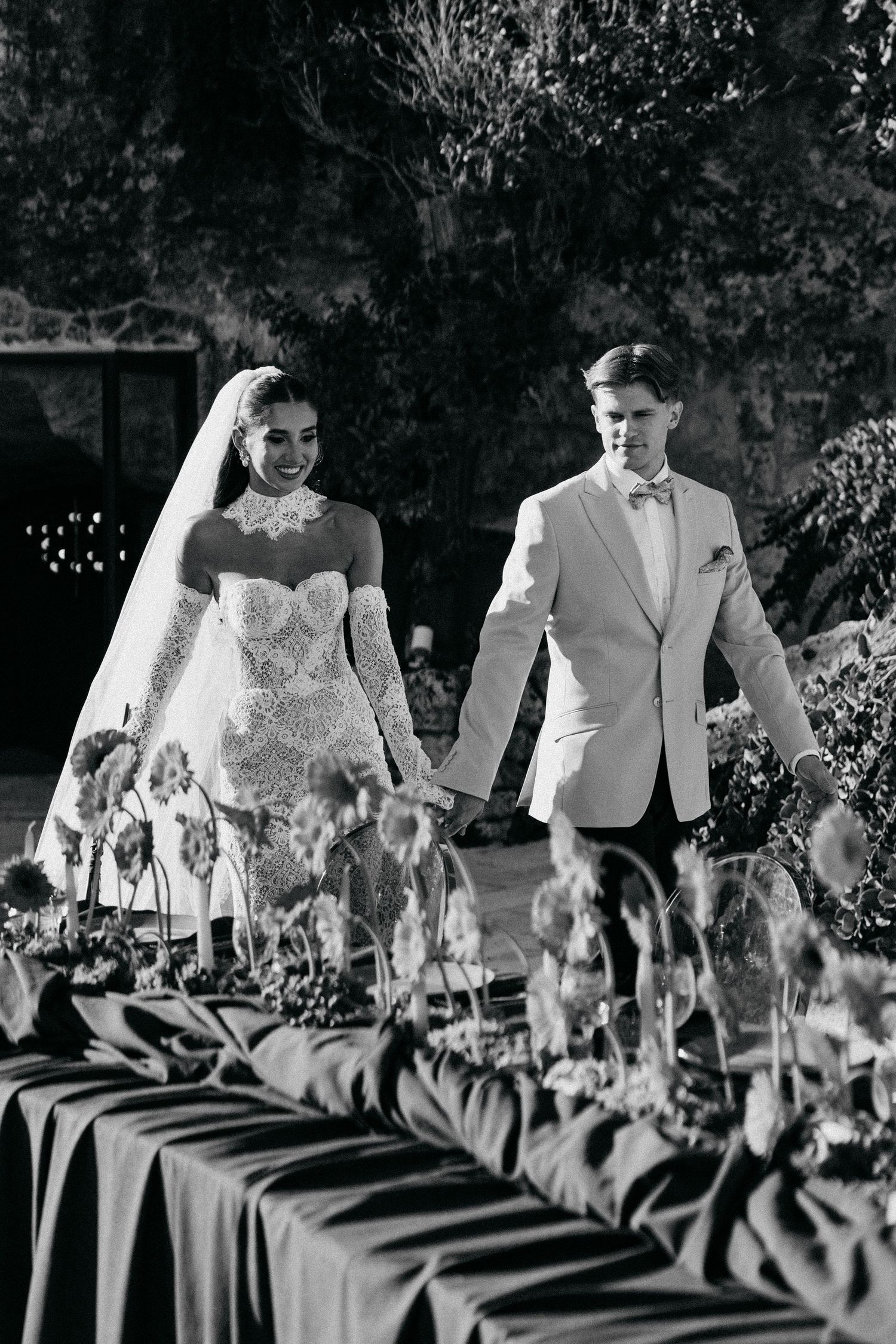 Black and white photo of a bride in a lace gown and a groom in a light suit holding hands beside a decorated wedding table.