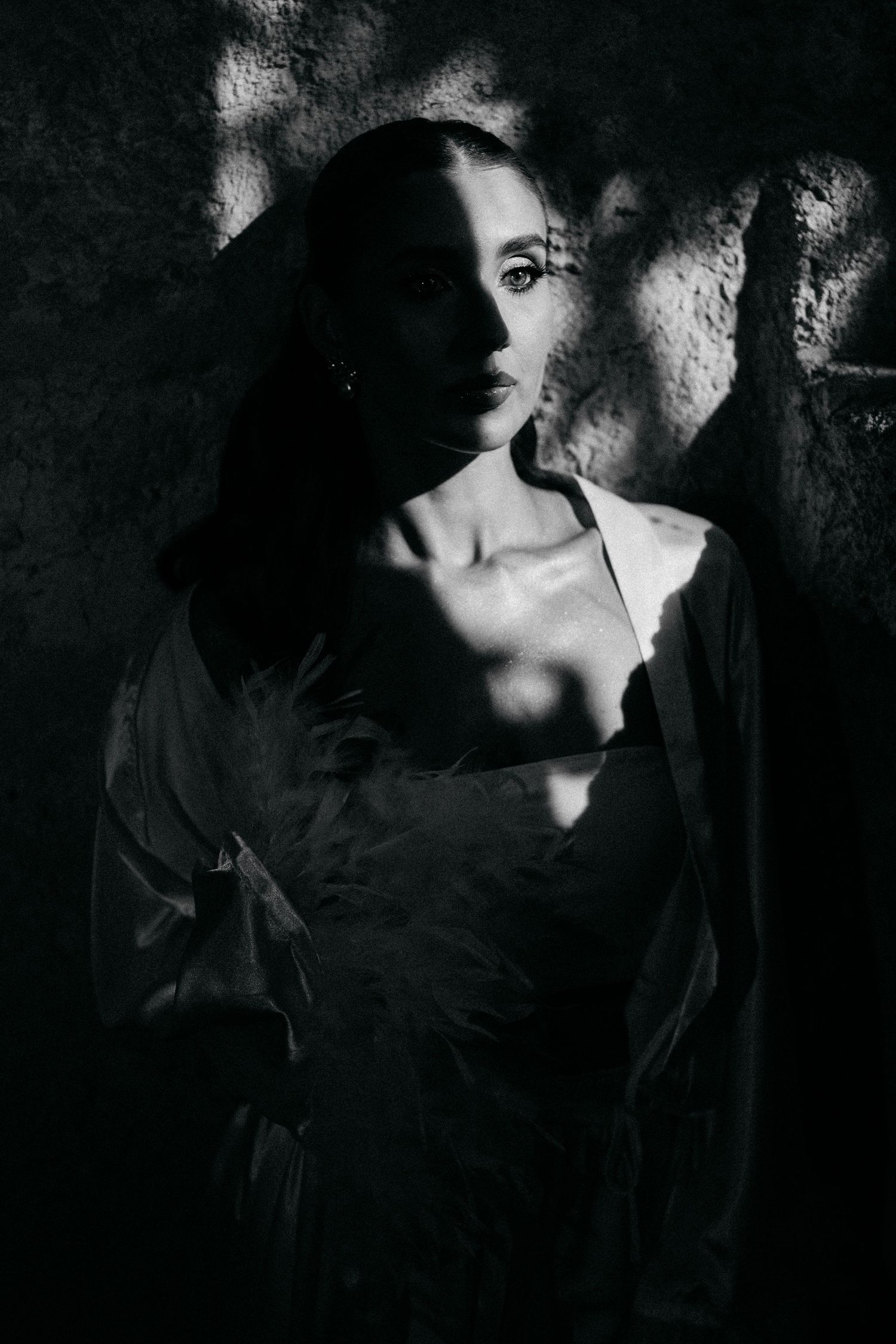 Black and white portrait of a woman with dark hair in a feathered robe, dramatic dappled light and shadow falling across her face.