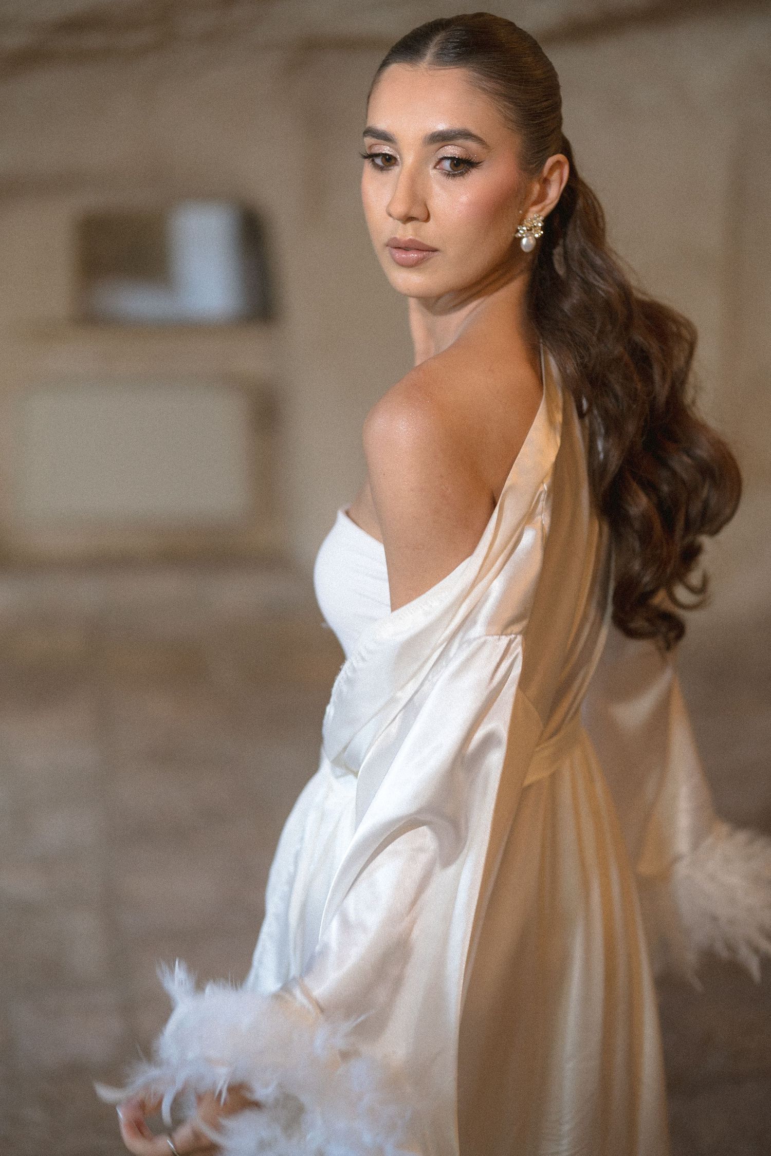 Bride in a white satin robe with feather-trimmed sleeves and sparkling earrings, glancing over her shoulder in a soft-lit stone interior.