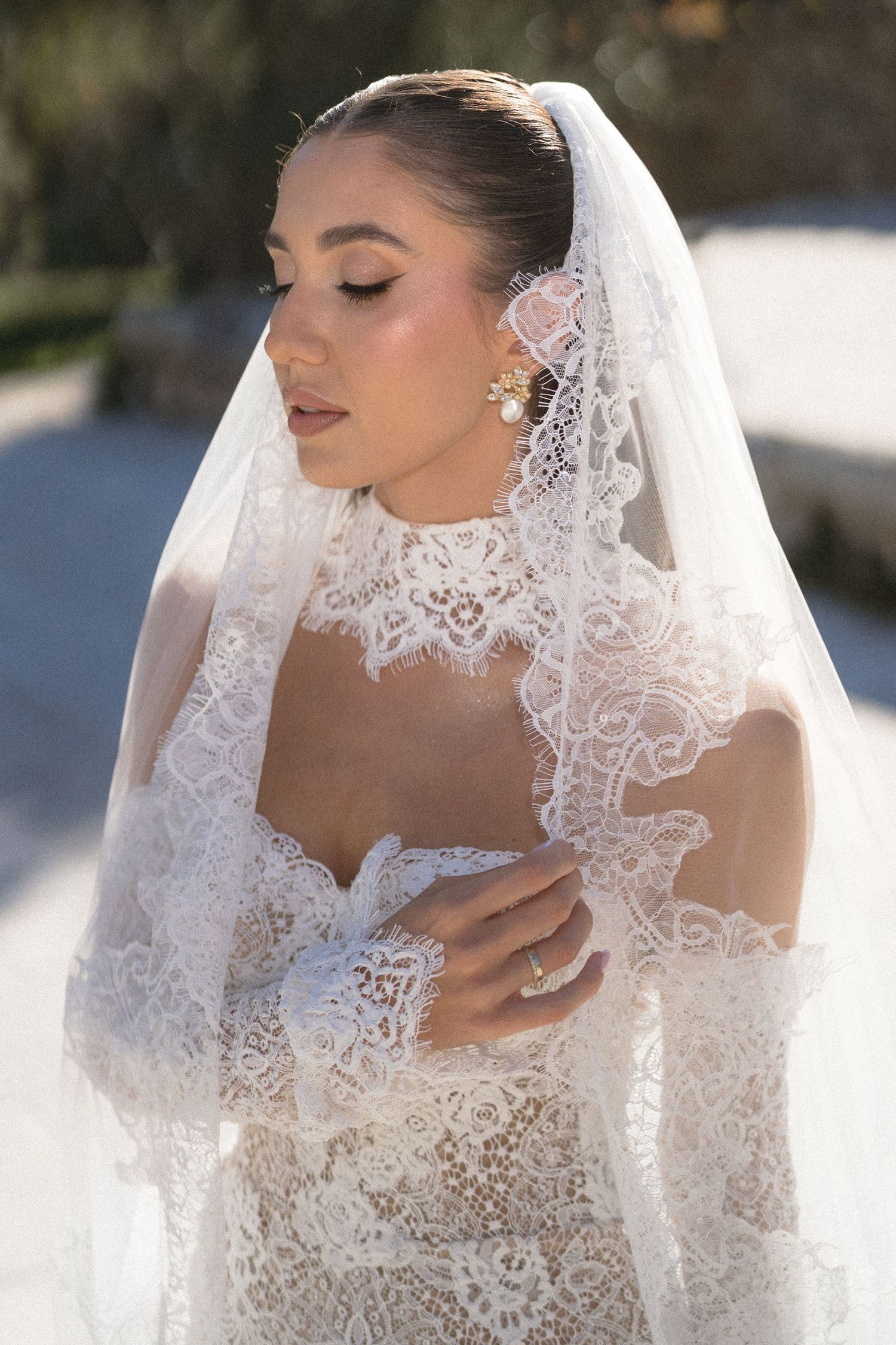 Bride in profile wearing a lace gown with high collar, a floral hair accessory, statement earrings, and a lace-trimmed veil.