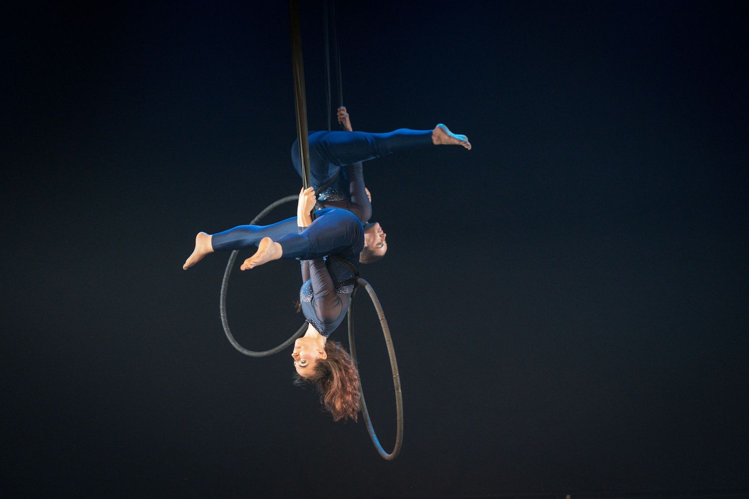 Aerial performer in blue costume gracefully poses on a suspended hoop against dark background.