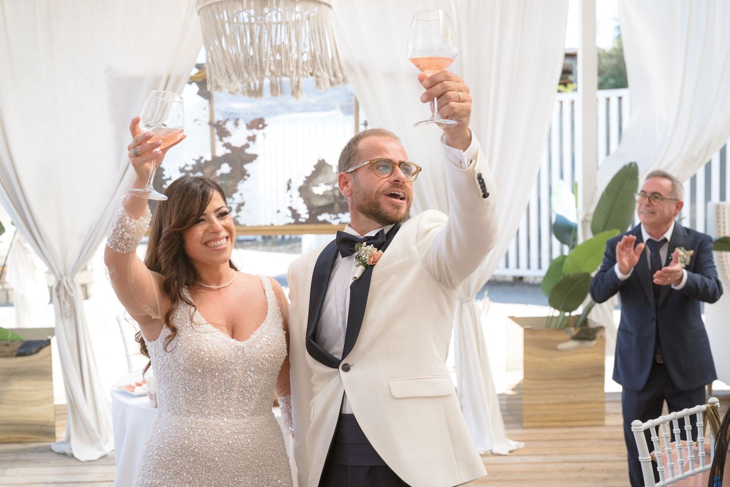 Newlyweds in white attire raise their glasses in celebration during wedding reception.