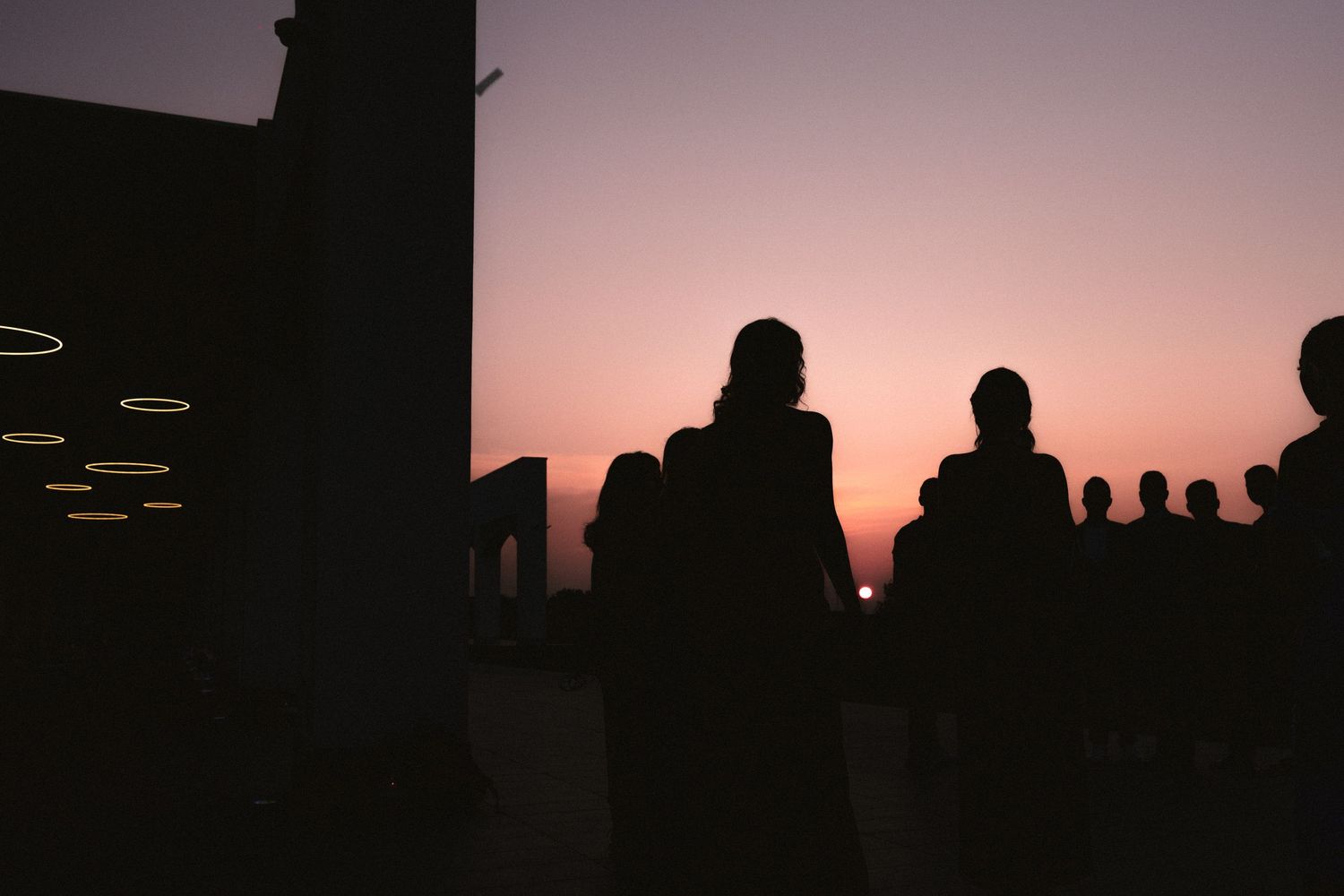 Silhouettes of people gathered on a rooftop terrace at sunset with a vibrant pink and purple sky backdrop.