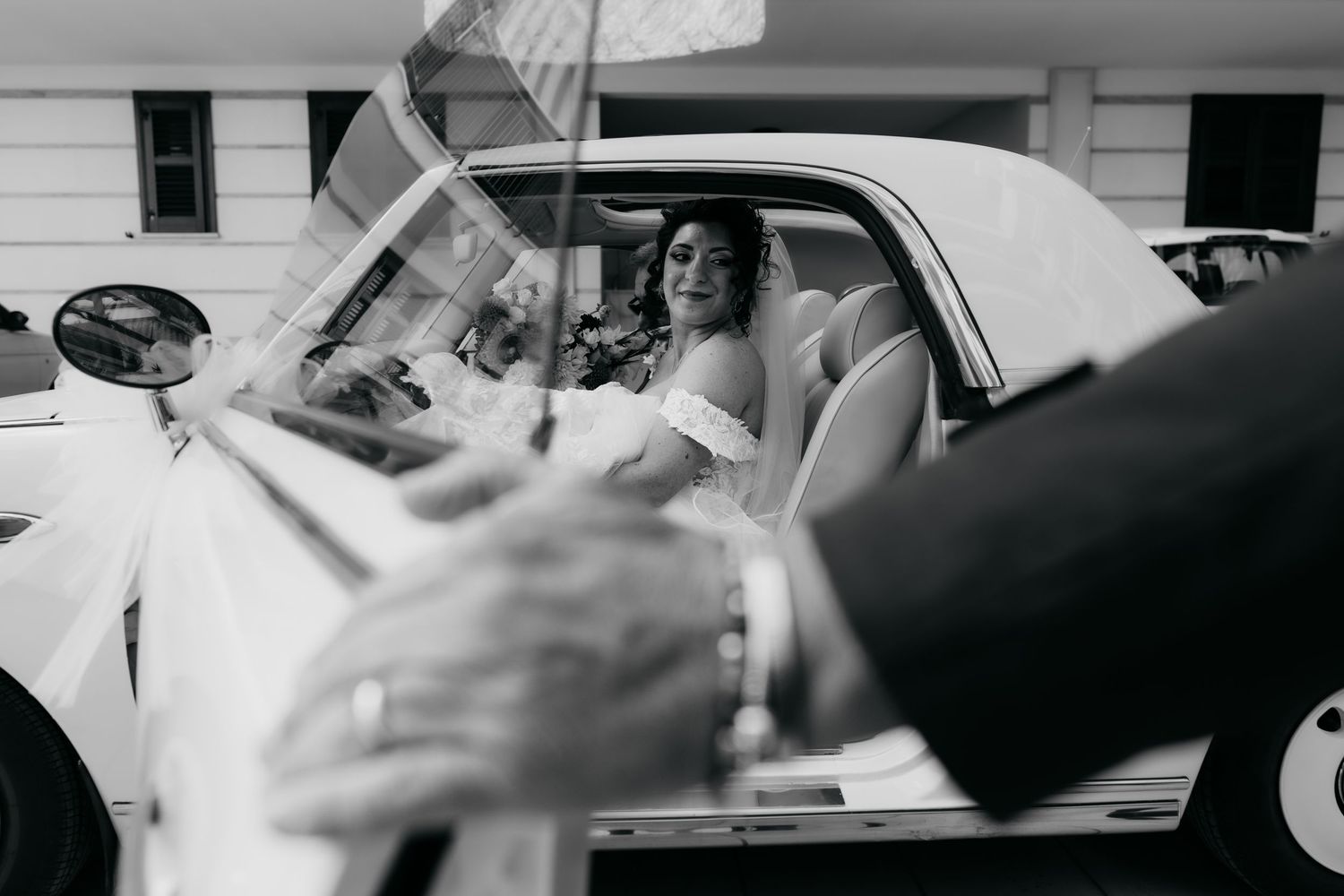 Bride in white dress sitting in a vintage white convertible car on her wedding day, black and white photo.