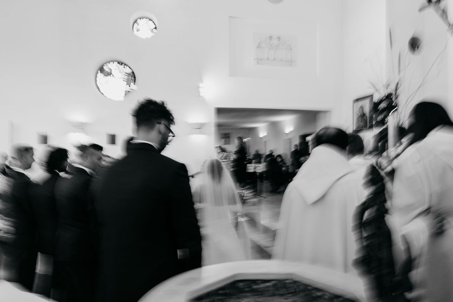 Black and white blurred motion photo of wedding guests and priest inside a church.