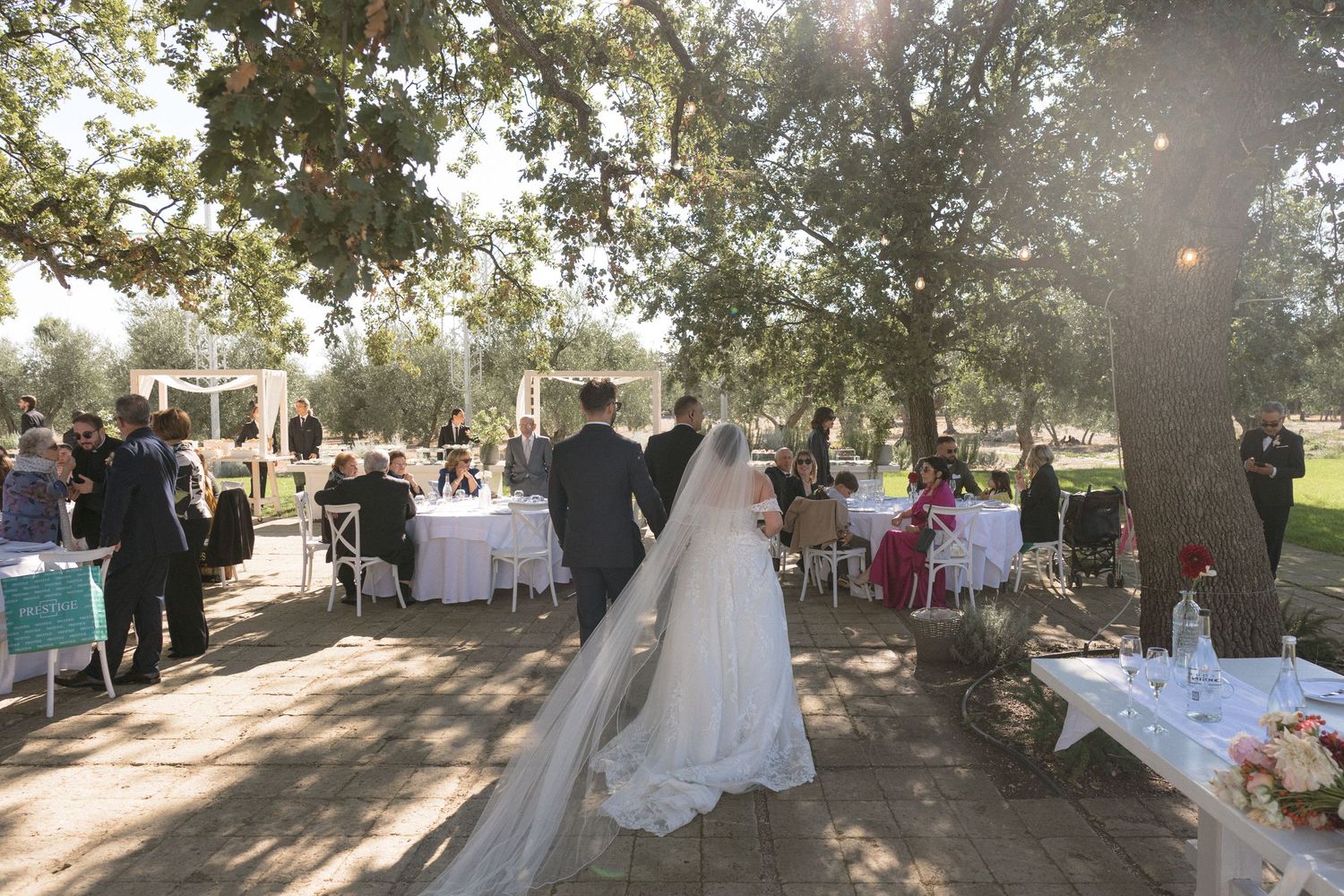 A bride and groom walk through an elegant outdoor wedding reception under large oak trees with white table settings.