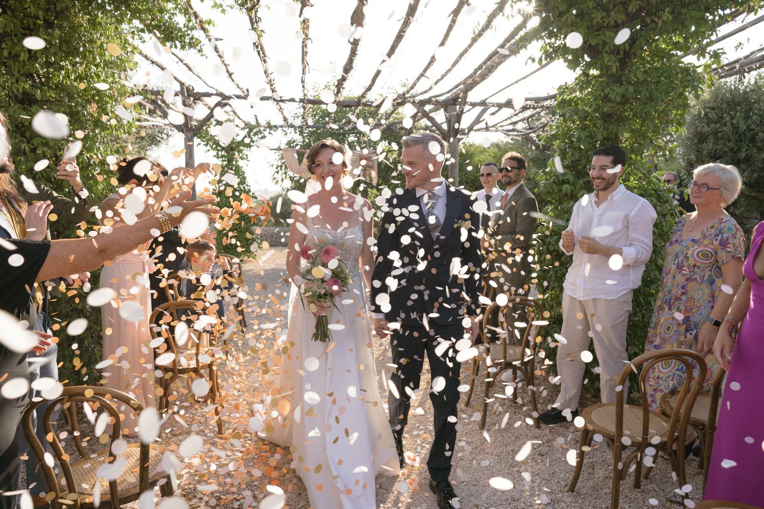 Bride and groom walk through confetti shower surrounded by guests at an outdoor wedding ceremony.