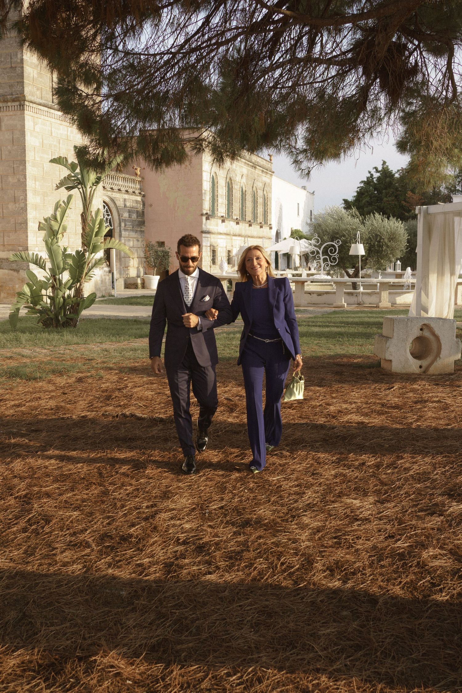 A couple walks together on a dirt path near a historic building during an outdoor wedding.