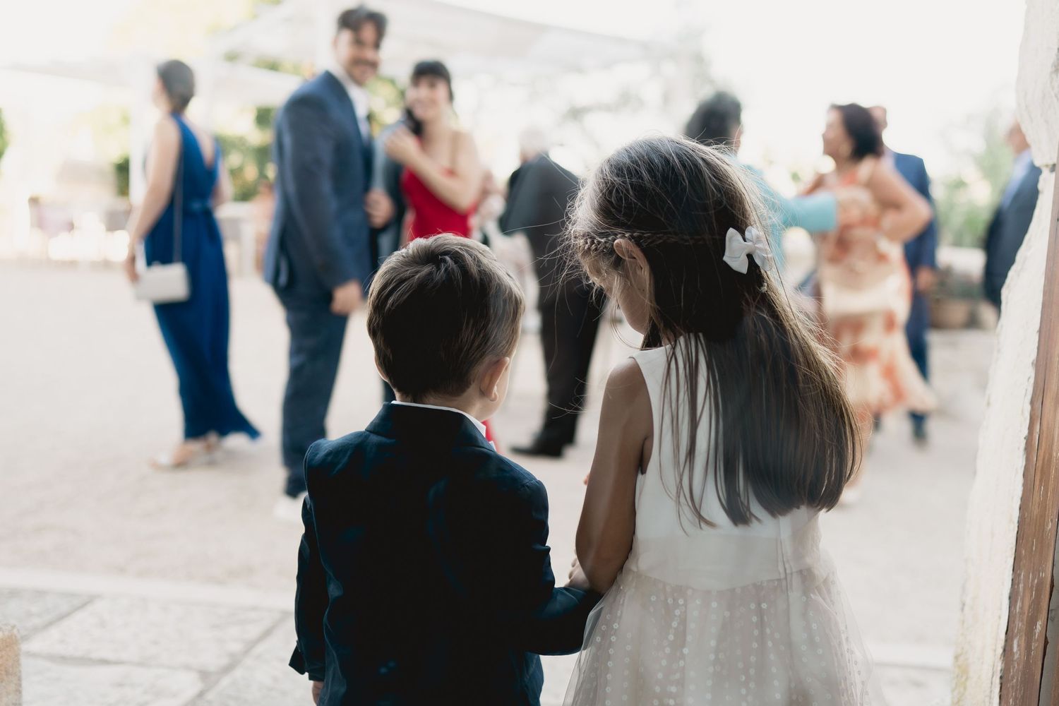 Two children watch a celebration indoors while standing together in formal attire.