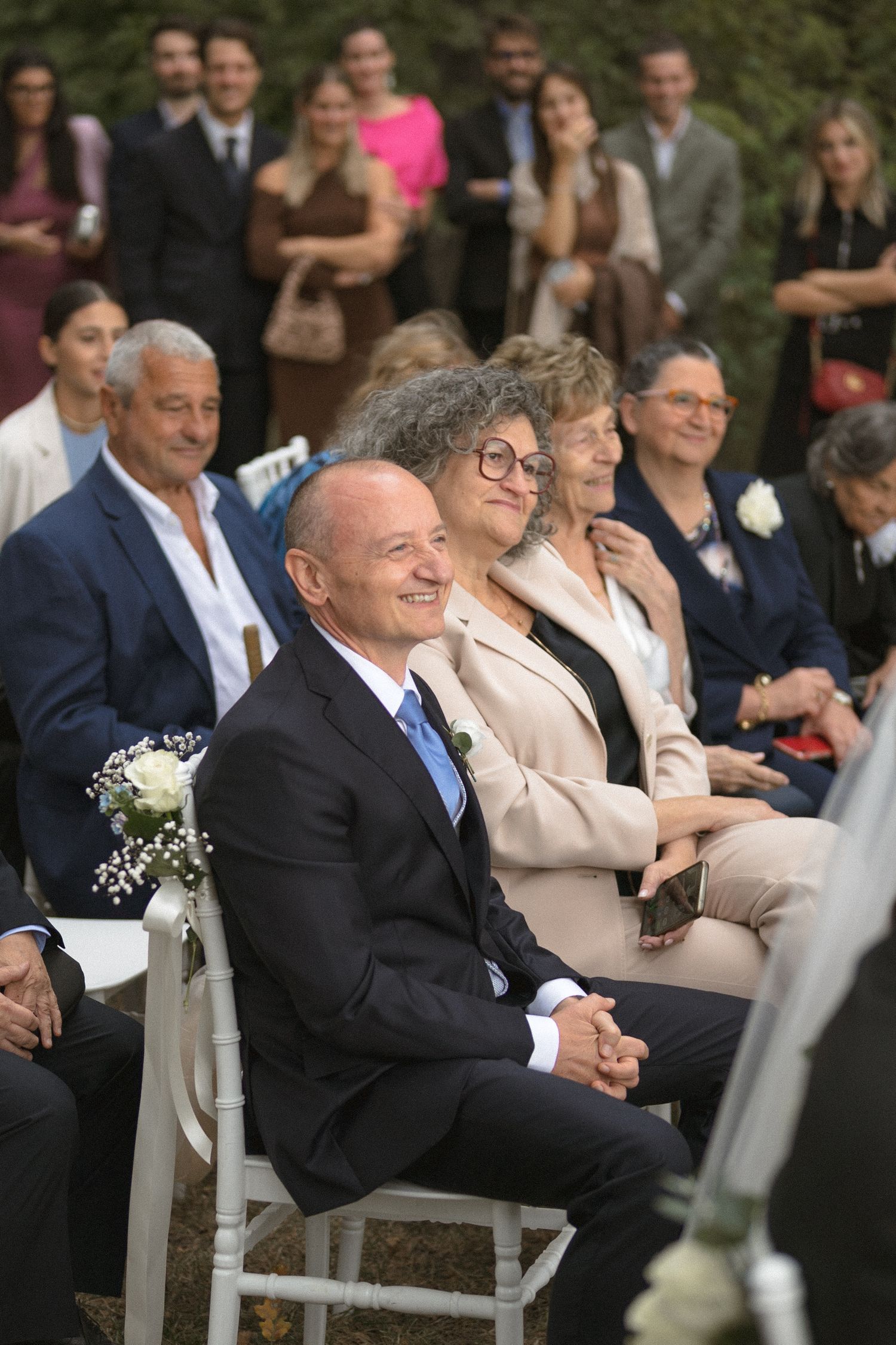 Wedding guests seated in formal attire watching ceremony outdoors.