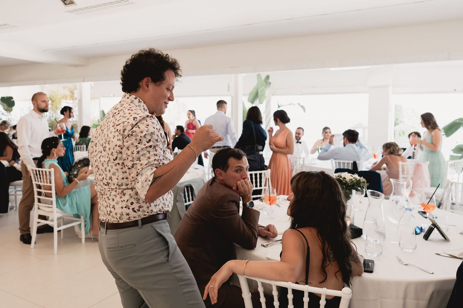 Wedding reception celebration with guests seated at white tables in a bright indoor venue.
