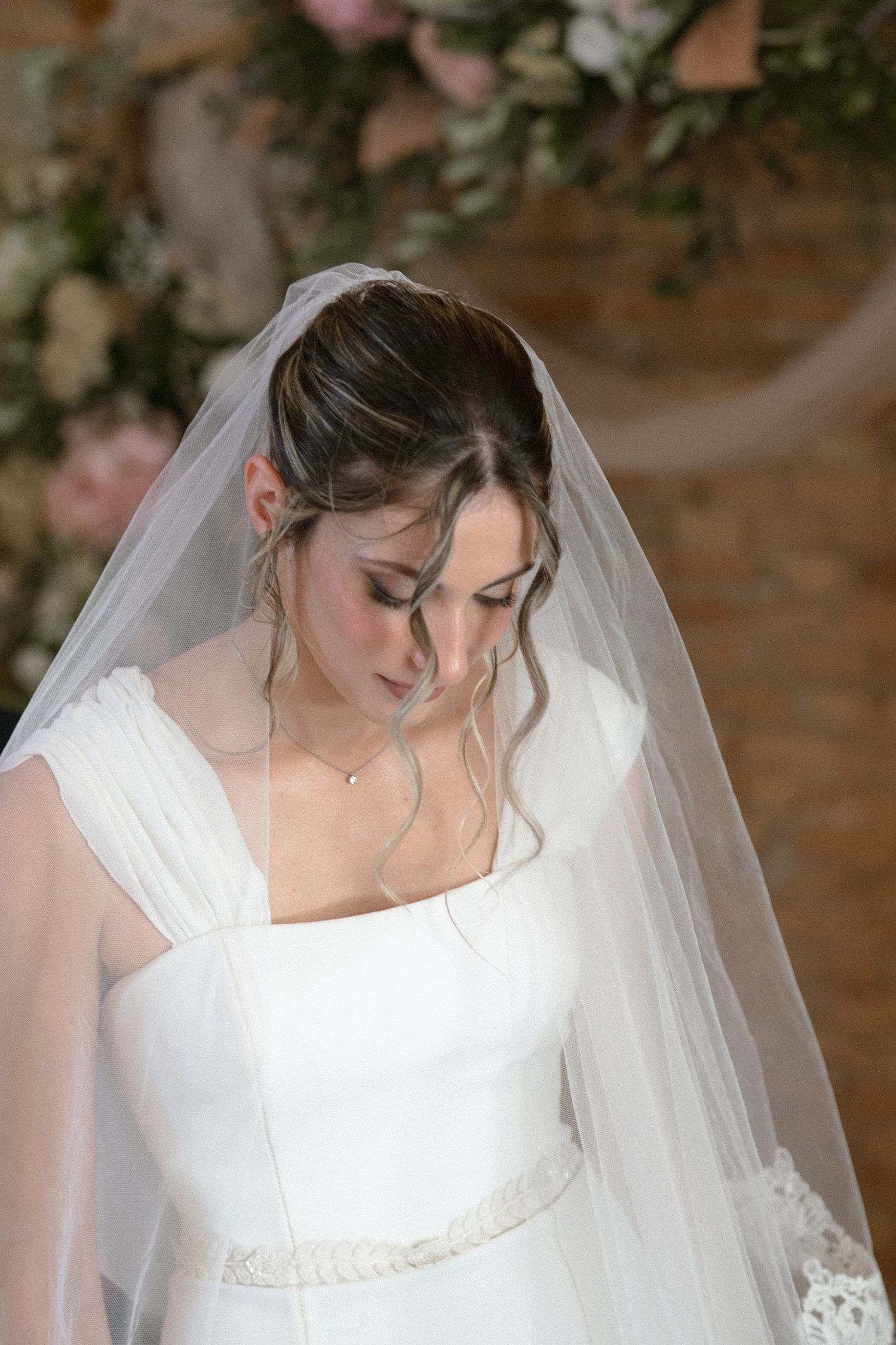 A bride wearing a classic white wedding dress with a flowing veil looks down thoughtfully against a floral backdrop.