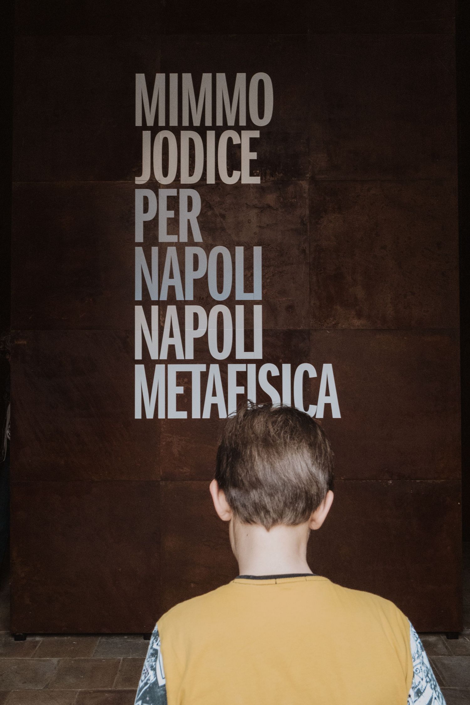 Person in yellow shirt stands in front of wall with Italian text 'Mimmo Iodice Per Napoli Napoli Metafisica'.