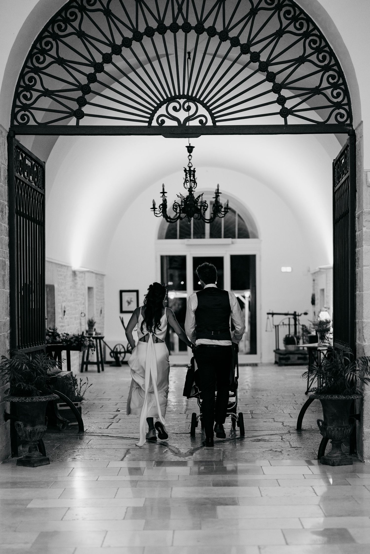 A couple walks through an elegant arched entrance with ornate ironwork and a chandelier overhead.
