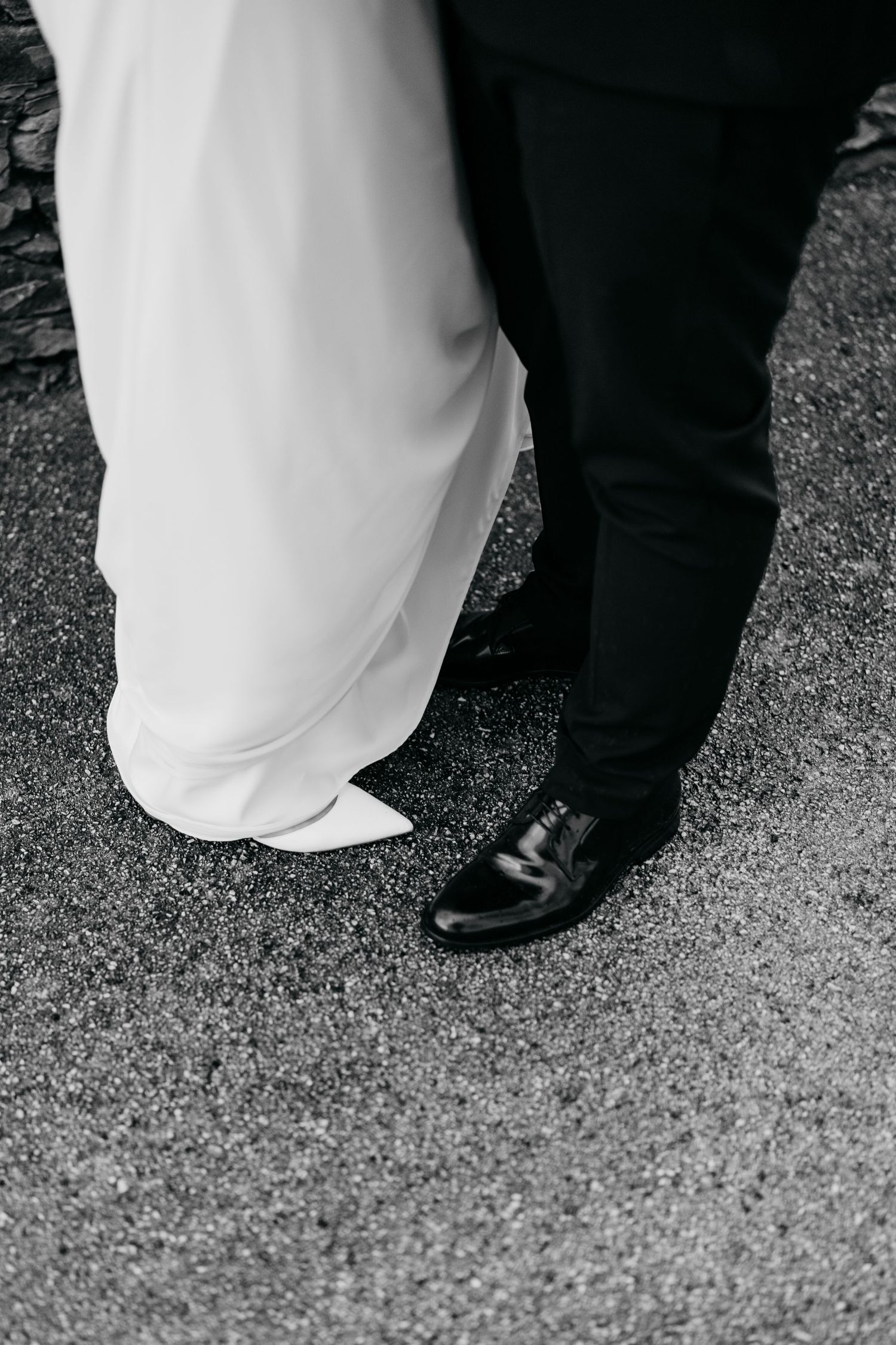 Black dress shoes and white wedding dress hem visible on a gravel path.