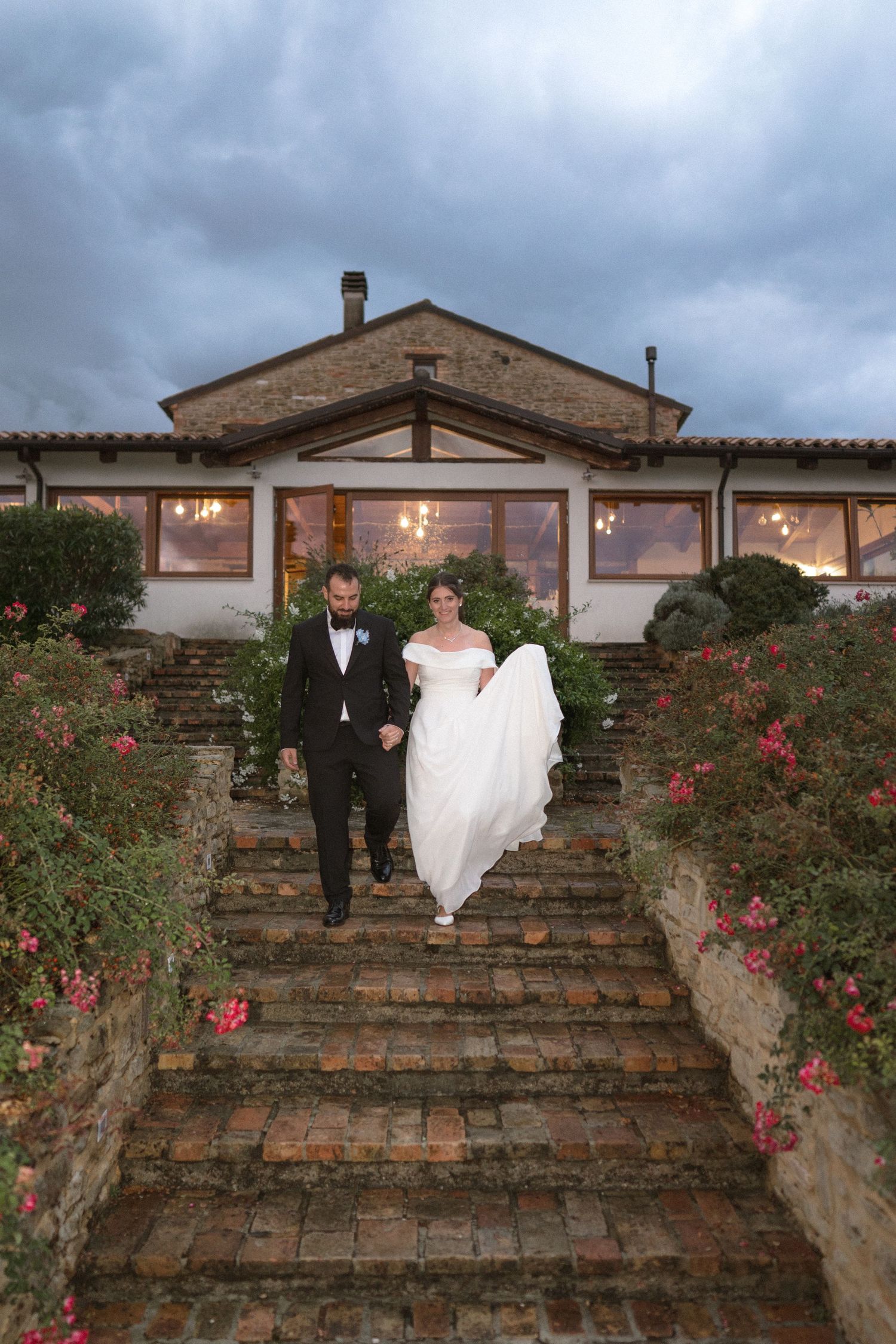 Wedding couple standing on stone steps with blooming red roses at dusk.