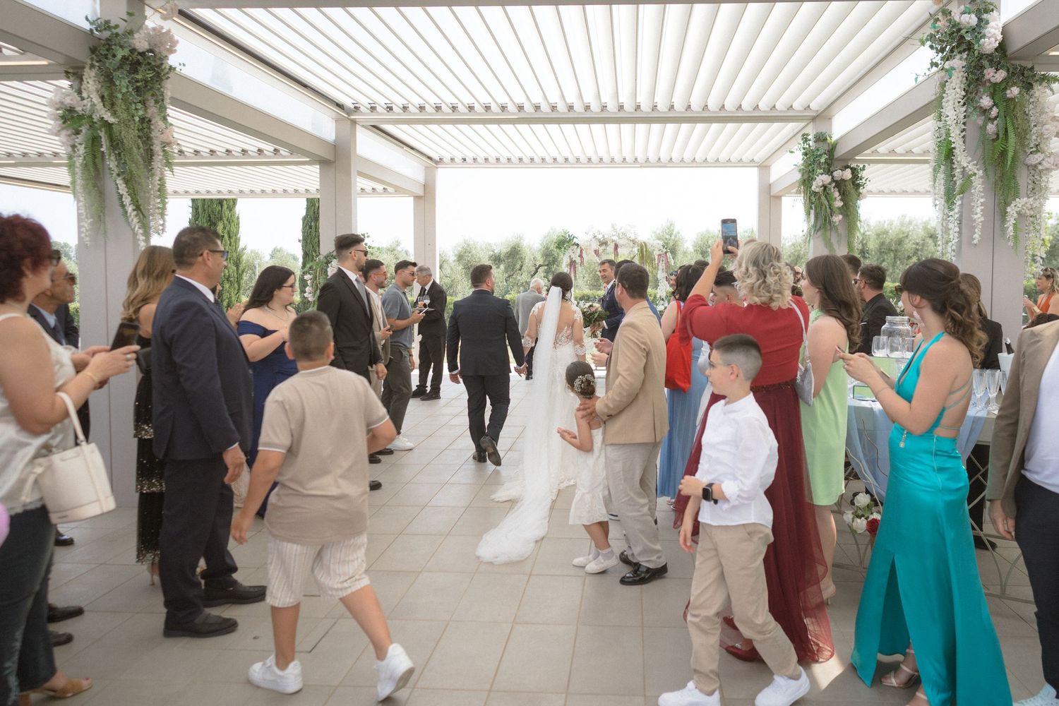 Wedding guests gather under an elegant white pergola decorated with greenery during an outdoor reception.