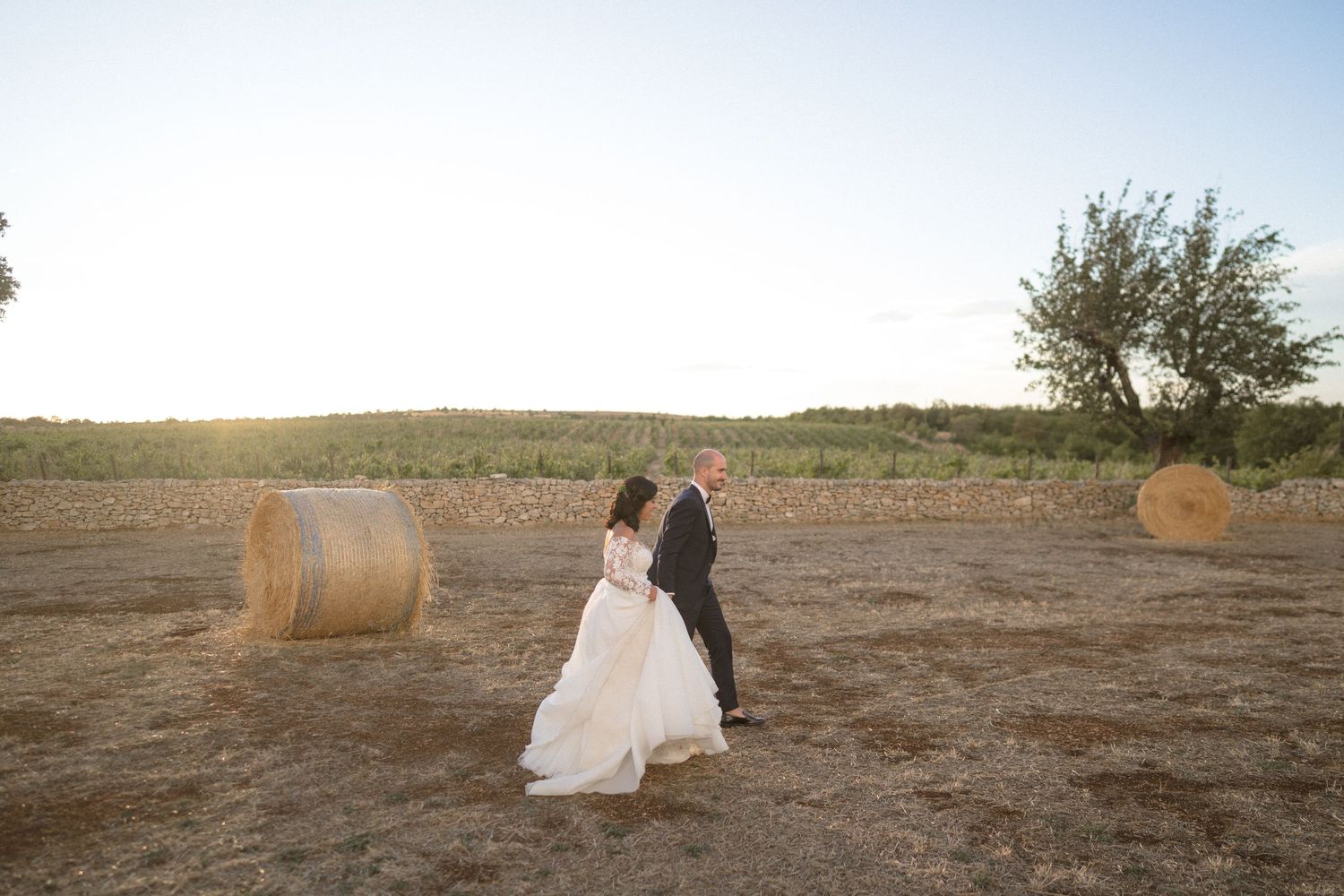 Wedding couple walking in a golden field at sunset with round hay bales in the background.