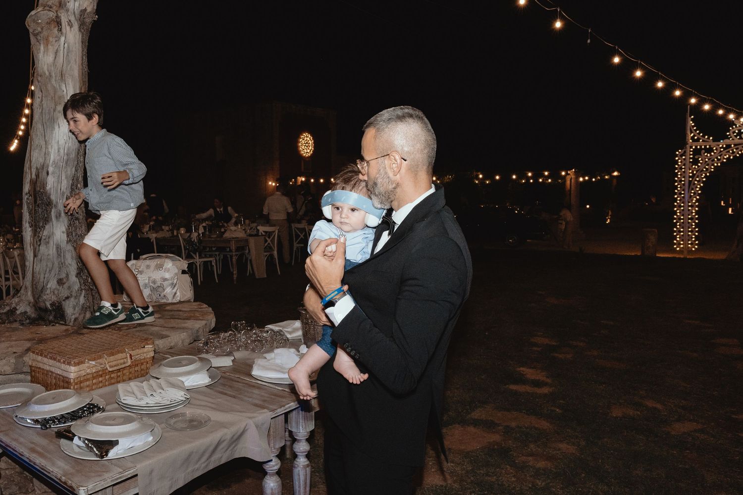 Wedding guests dance and celebrate under twinkling string lights at an outdoor evening reception.