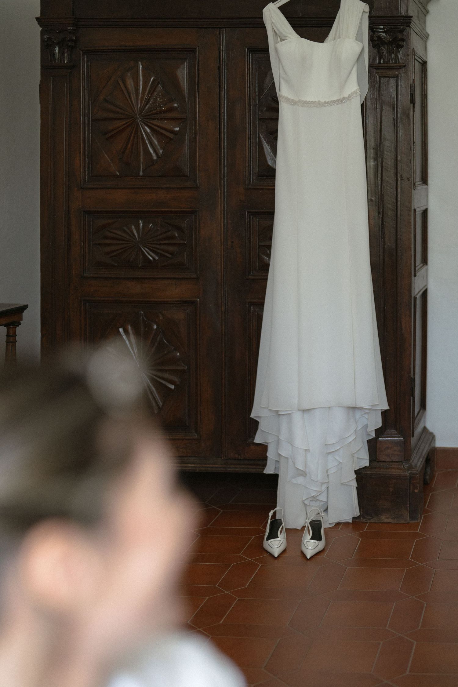 A white wedding dress hangs elegantly against a dark wooden door with decorative panels.