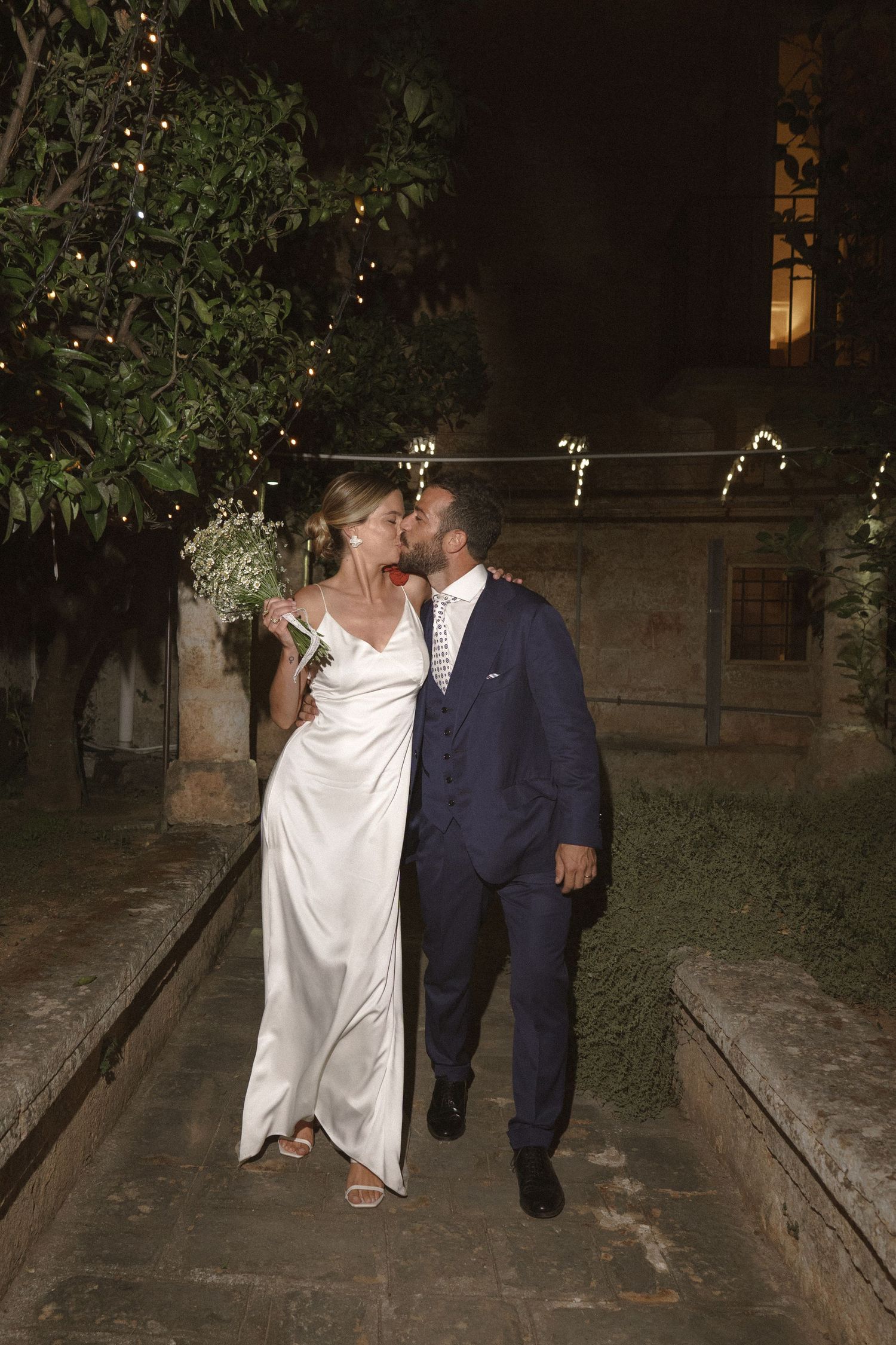 A romantic nighttime portrait with string lights and stone architecture in the background.
