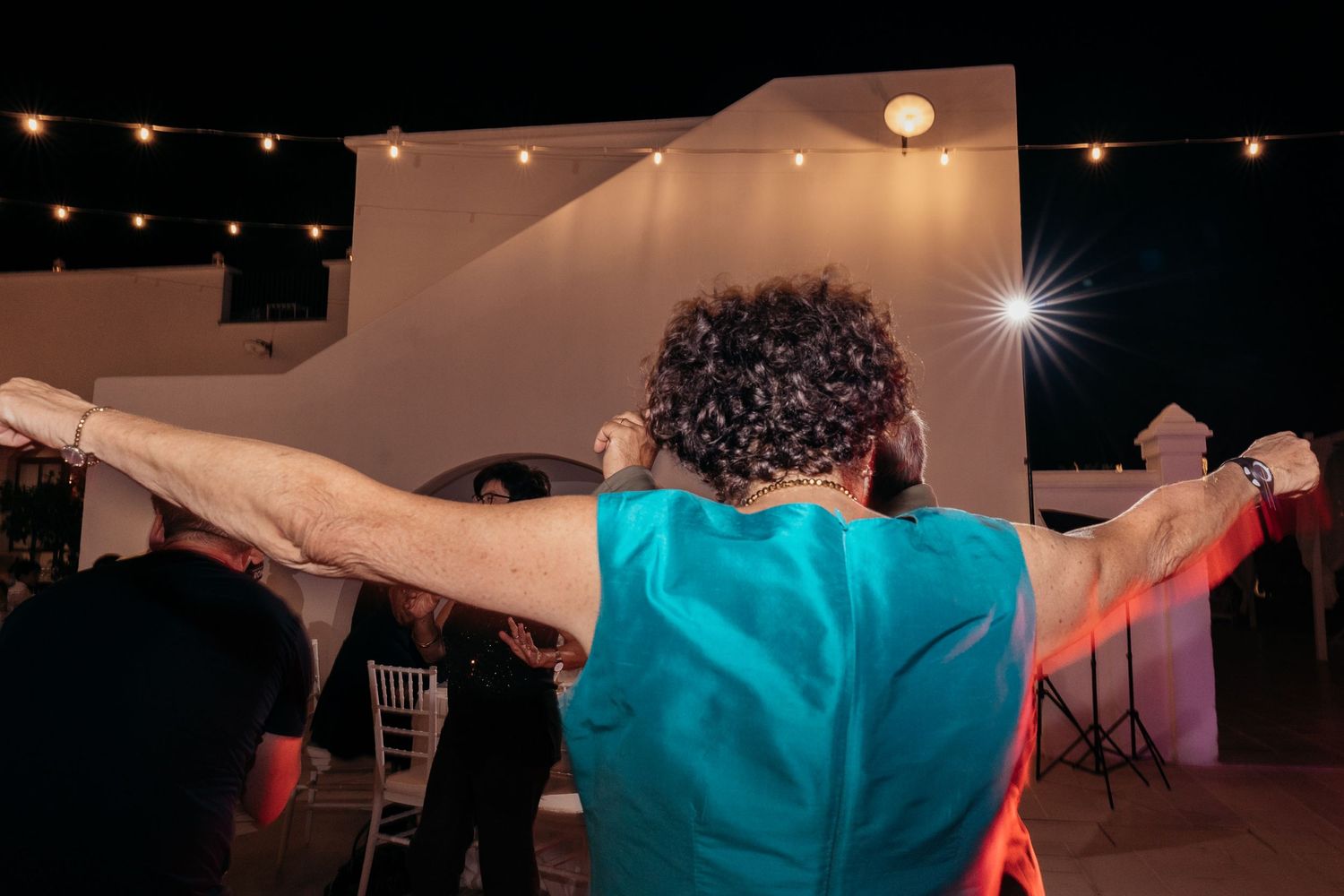 A person in a turquoise shirt dances energetically under outdoor string lights at a wedding reception.