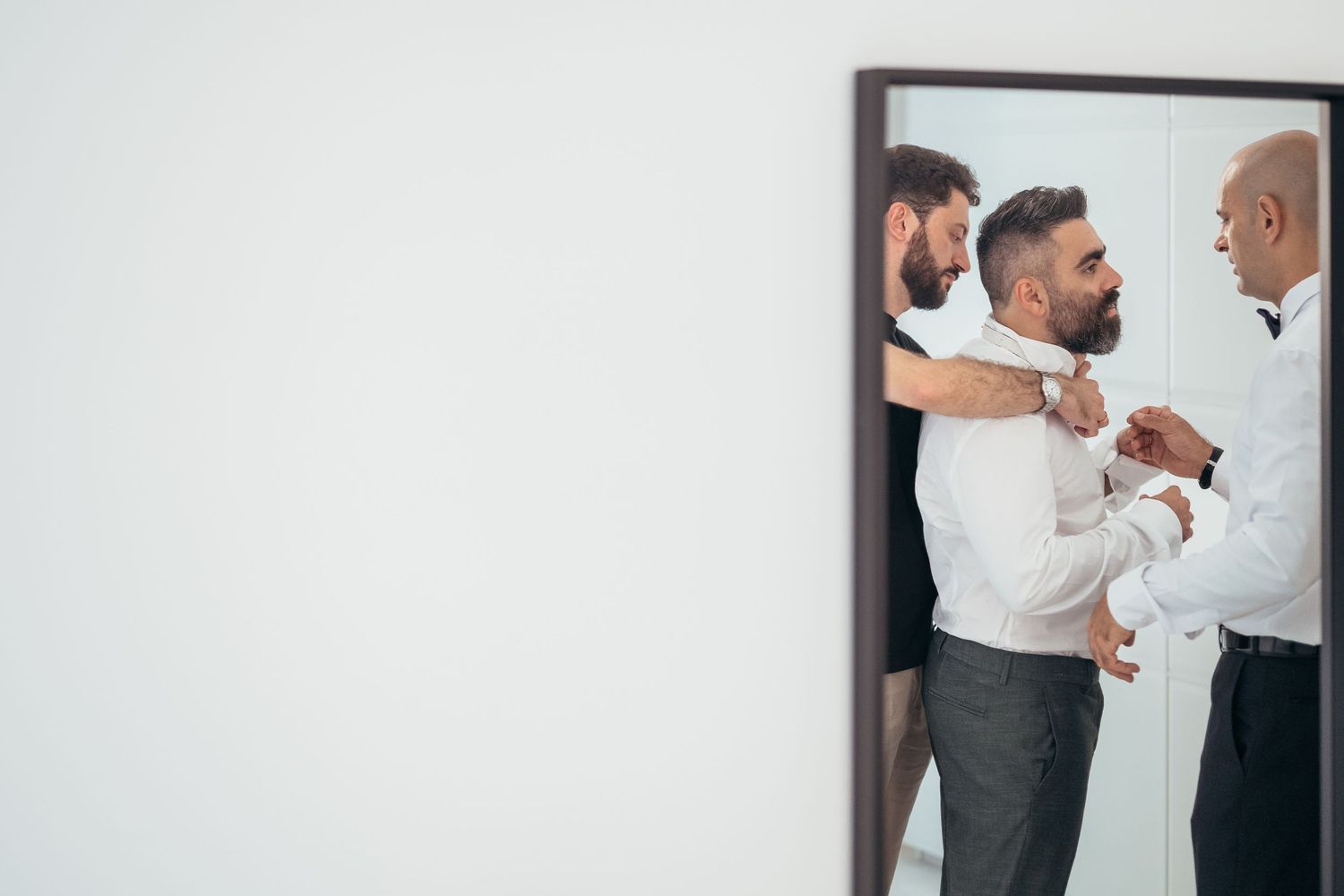 Mirror reflection shows people helping with formal attire adjustments during wedding preparations.