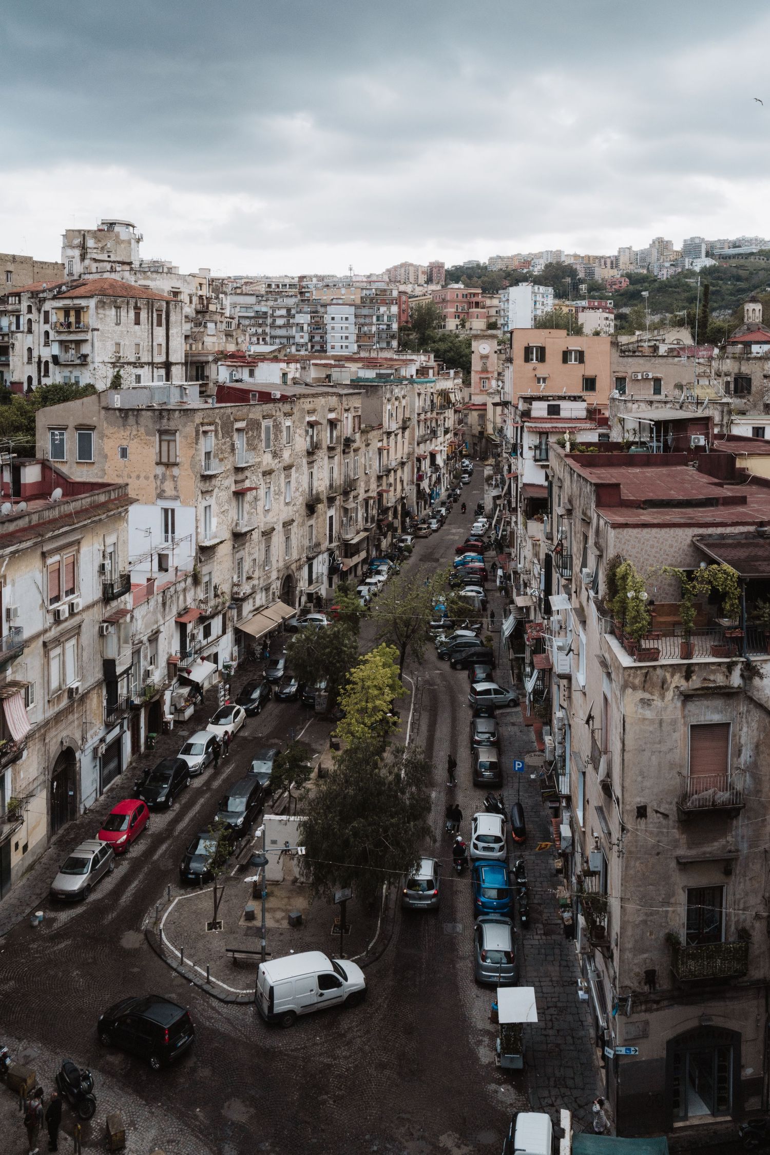 Aerial view of a narrow Italian street lined with historic buildings and parked cars under an overcast sky.