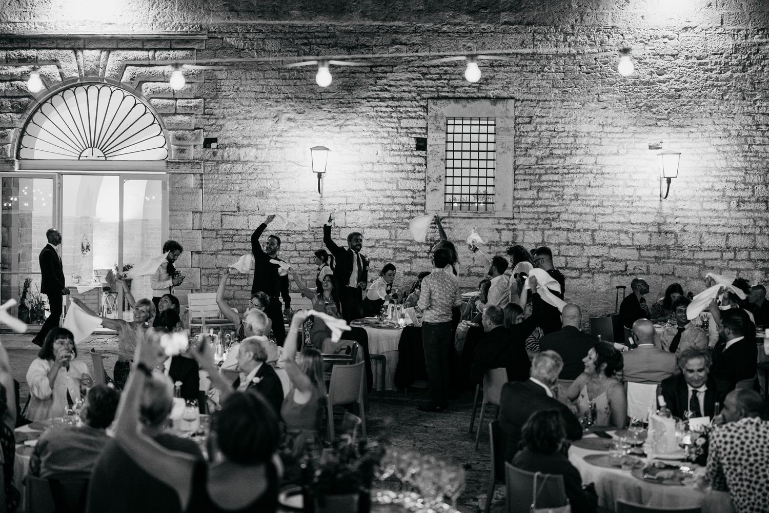 Wedding guests dance and celebrate in a rustic brick-walled venue with vintage lighting.