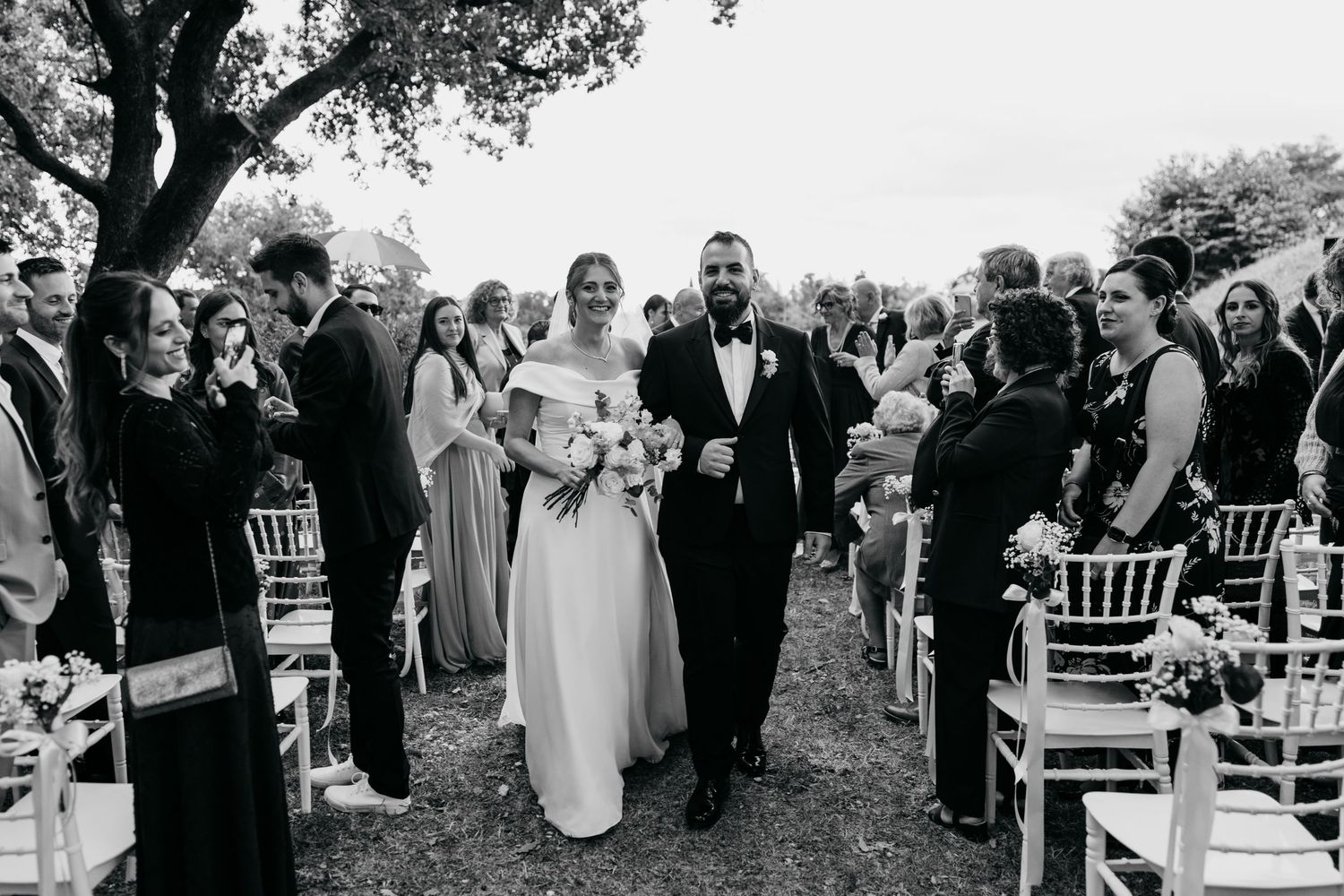 Black and white photo of wedding guests gathering for an outdoor ceremony among trees.