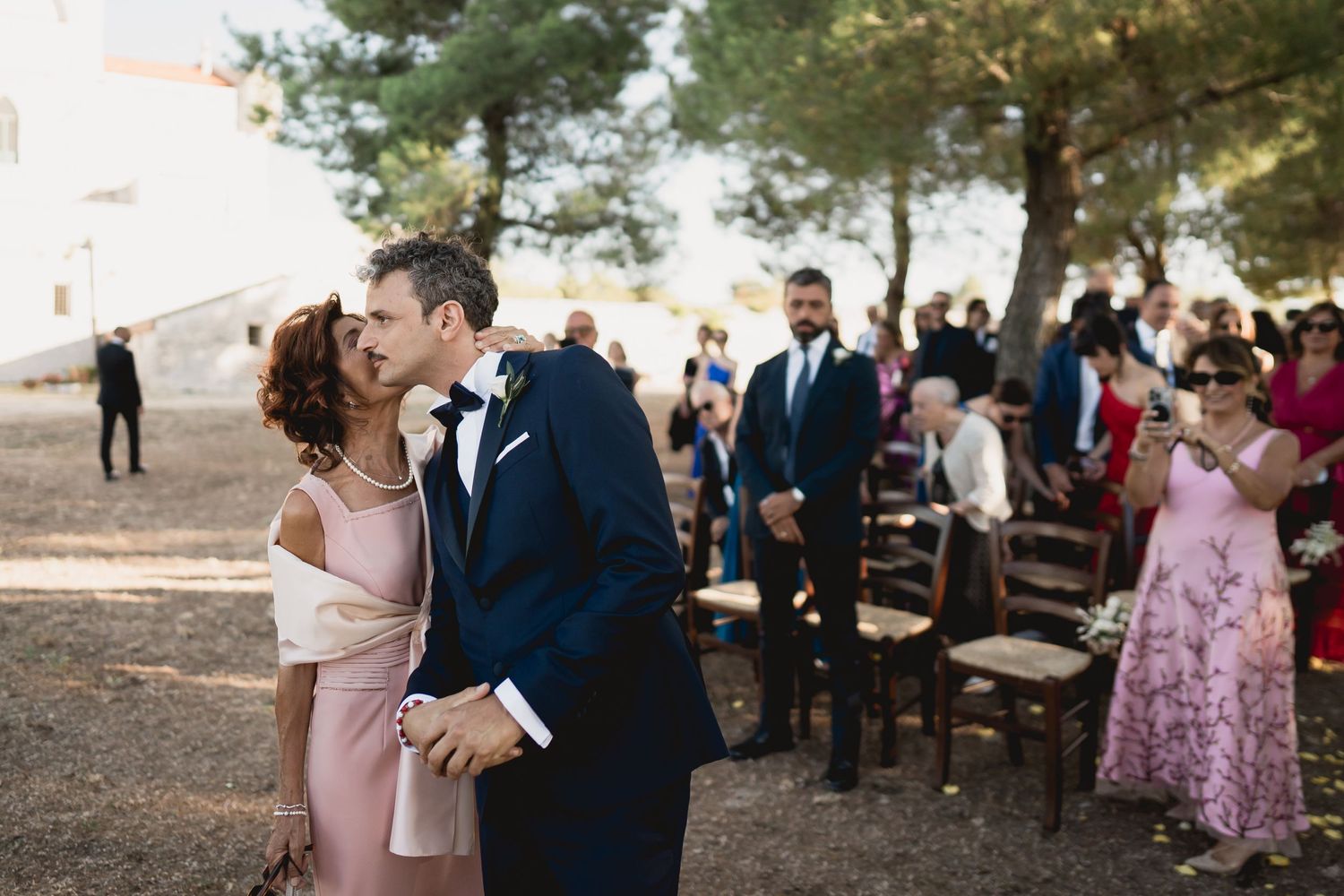 A couple embraces during an intimate outdoor forest wedding ceremony with guests seated on wooden chairs in the background.